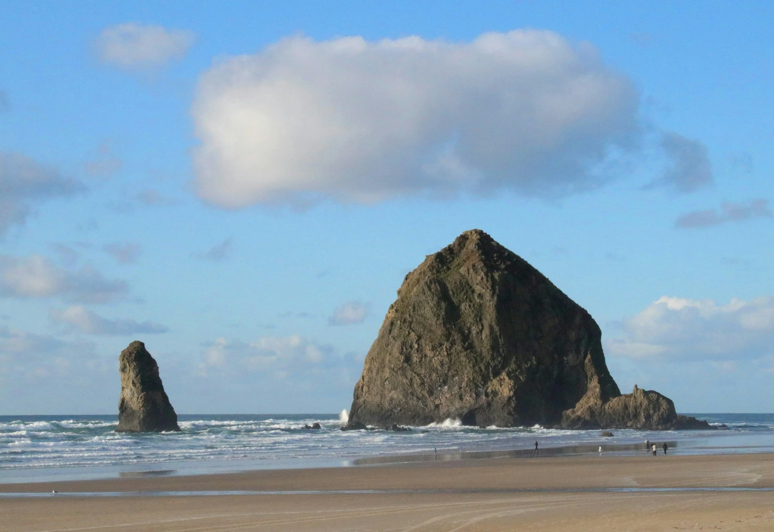 A coastal scene with large rock formations emerging from the ocean near a sandy beach, under a partly cloudy sky.