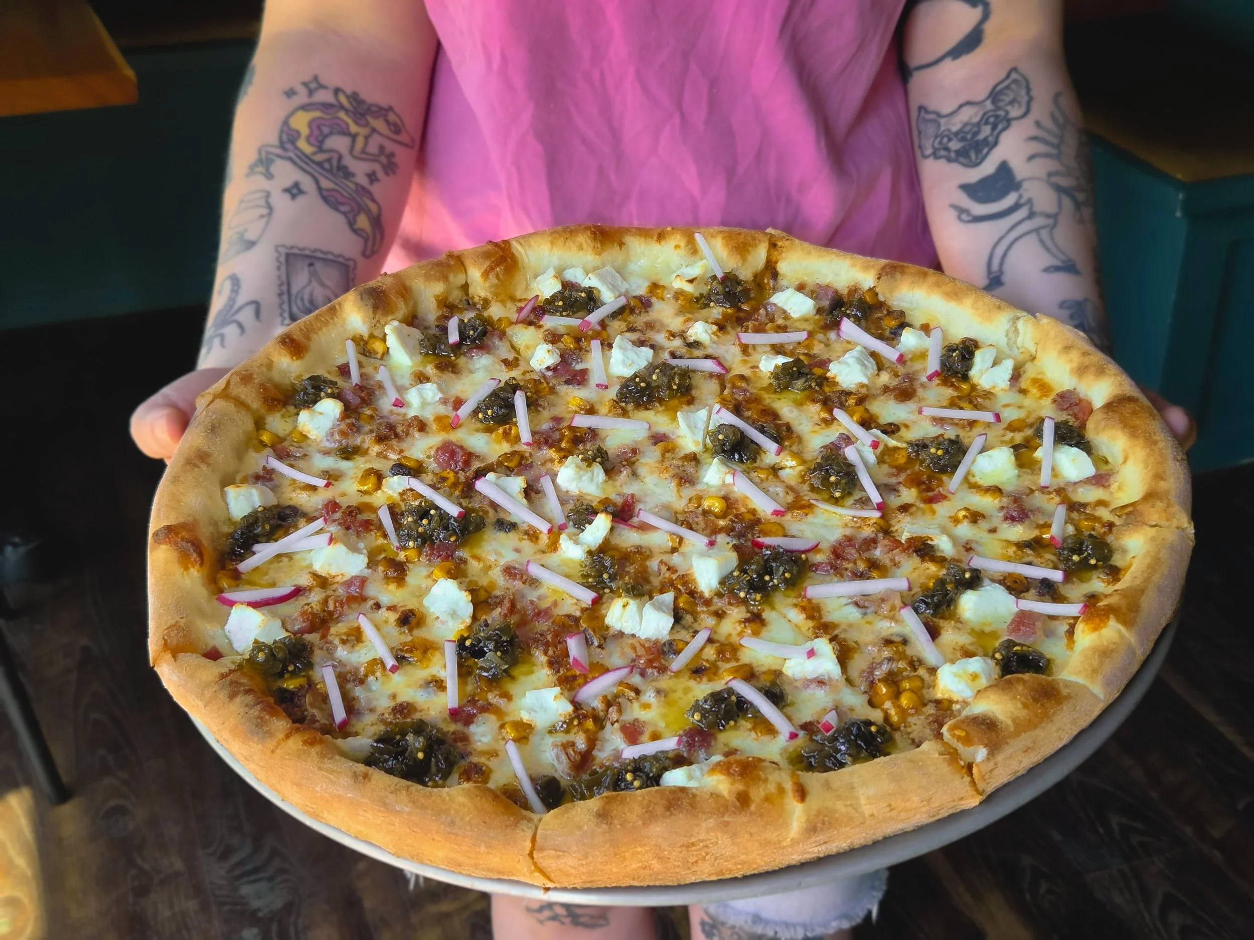 Person holding a freshly baked pizza with cheese, bacon, spiced corn, salata ricotta, green chili jam, and fresh sliced pink radish