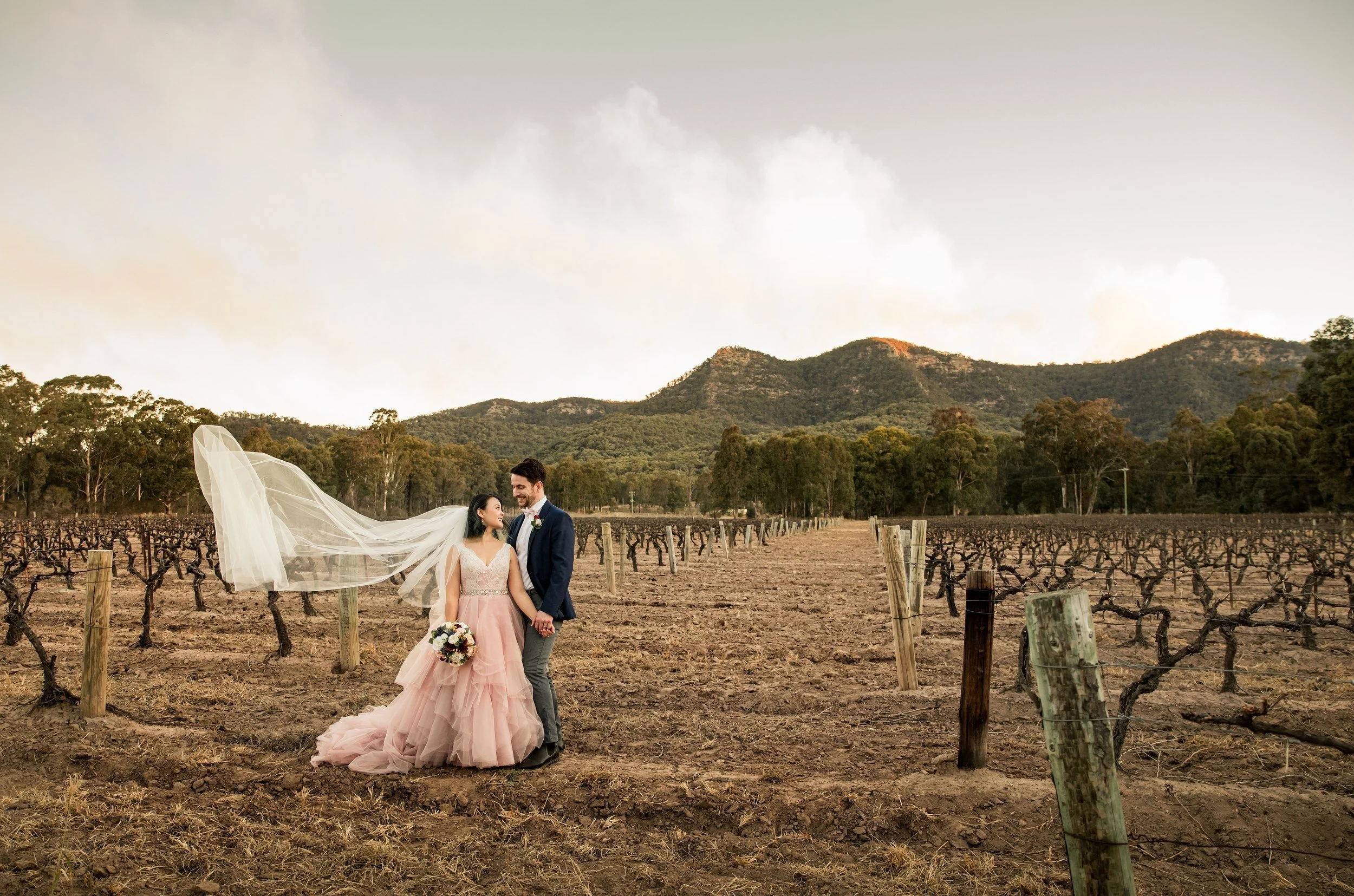 Couple standing in a winter vineyard with her veil flying in the breeze and a range of hills behind