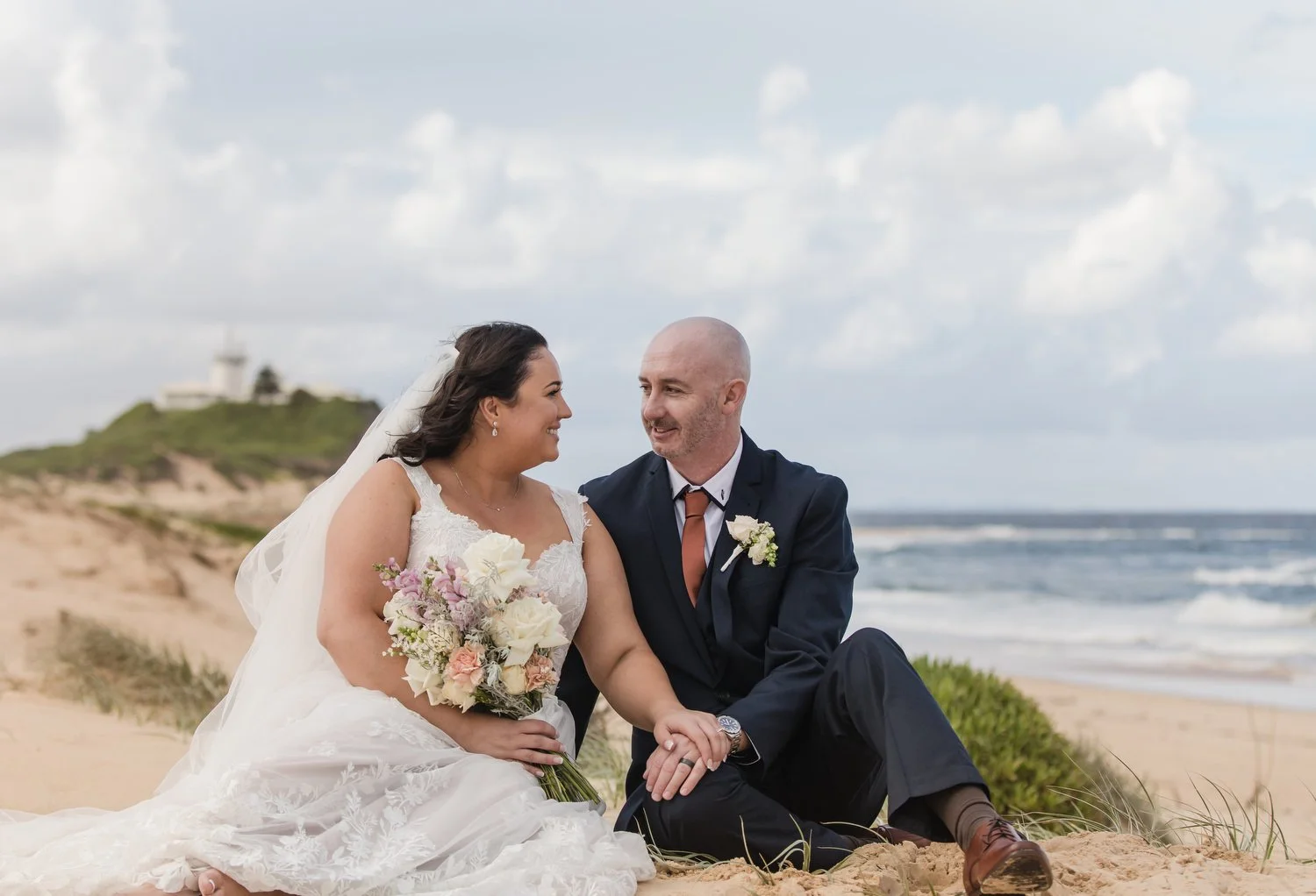 Bride and groom laughing in the sand dunes with the ocean behind