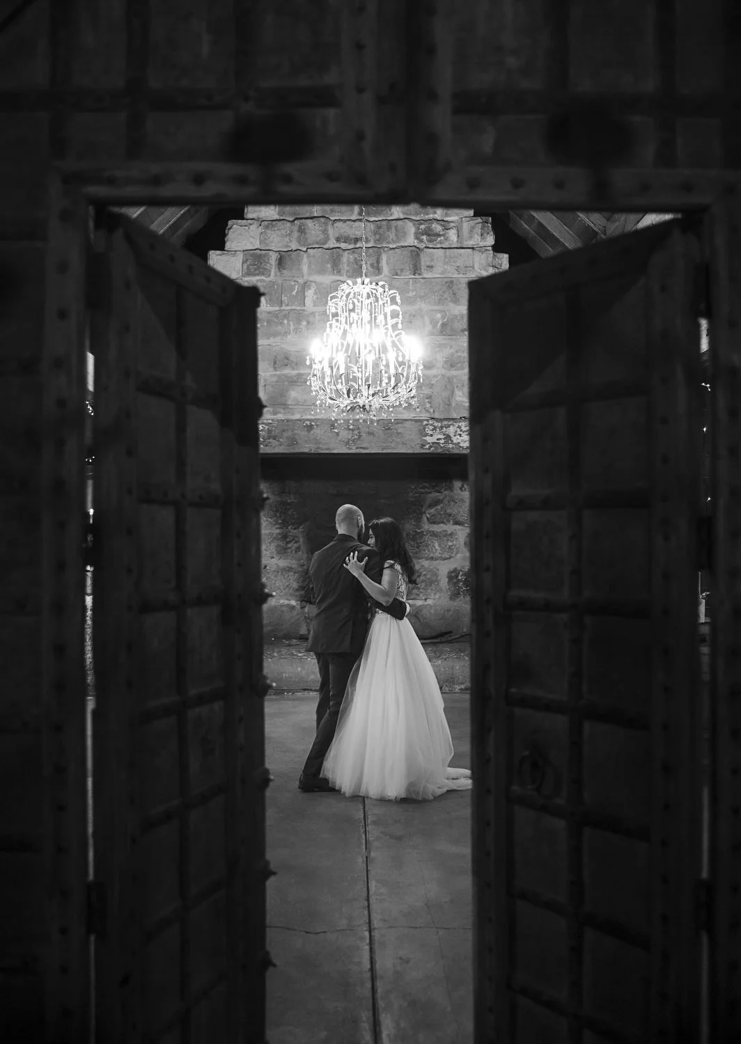 black and white image peeping through wooden doors of Peppers Creek Barrel Room  with Bride and Groom dancing 