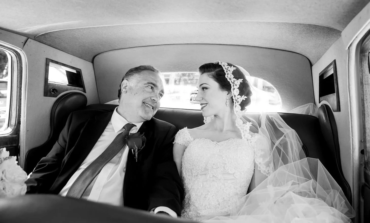 Bride in a lace wedding dress and veil sits smiling next to an older man in a suit with a boutonniere, inside a car.