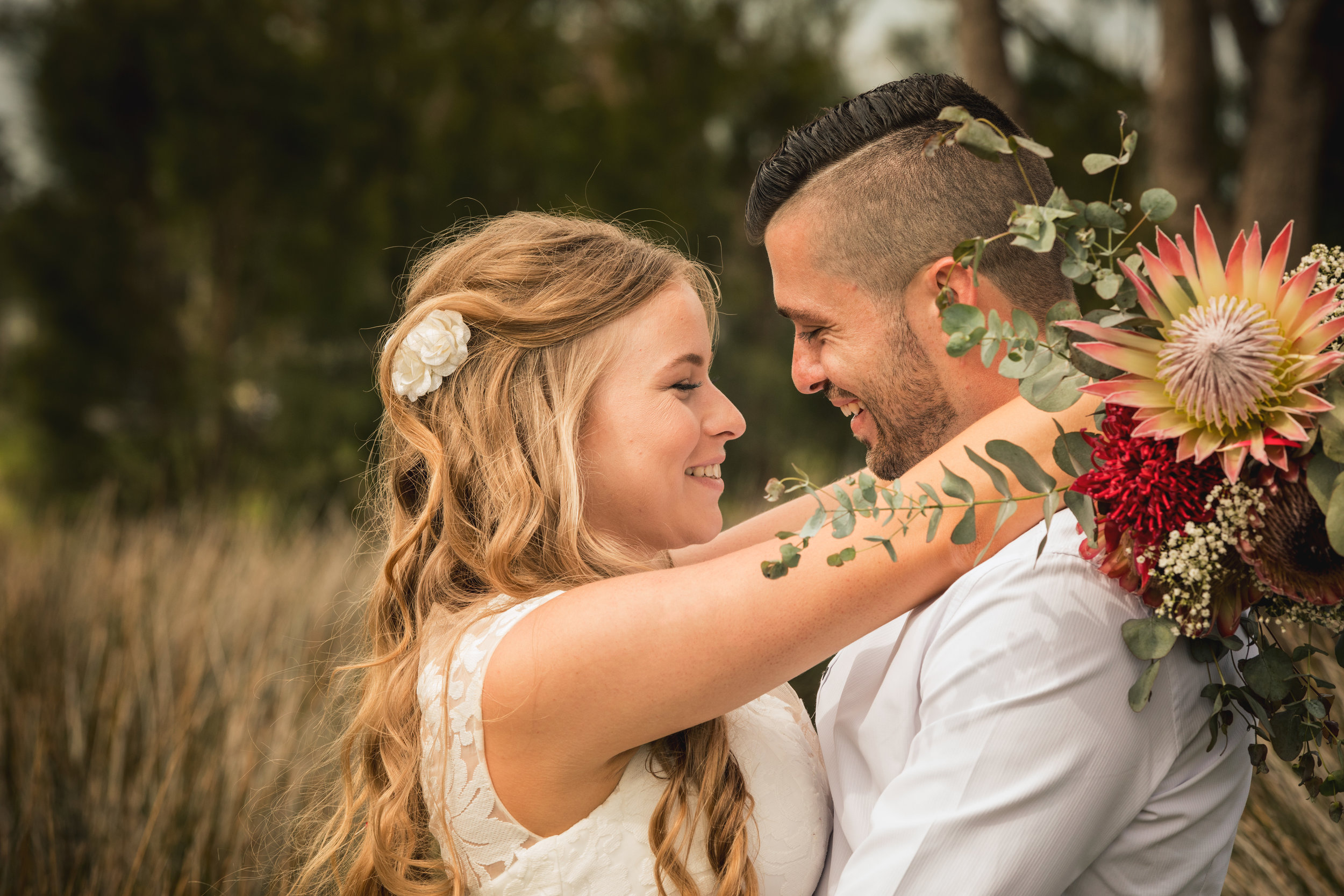 A couple embracing outdoors with a bouquet, smiling at each other, wearing wedding attire.