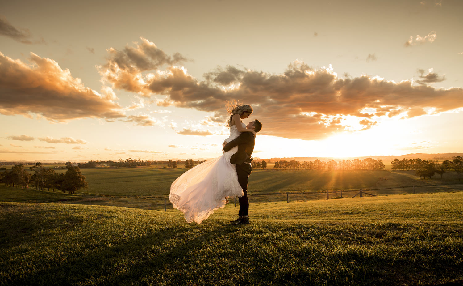 Couple in wedding attire in a field at sunset, groom lifting bride.