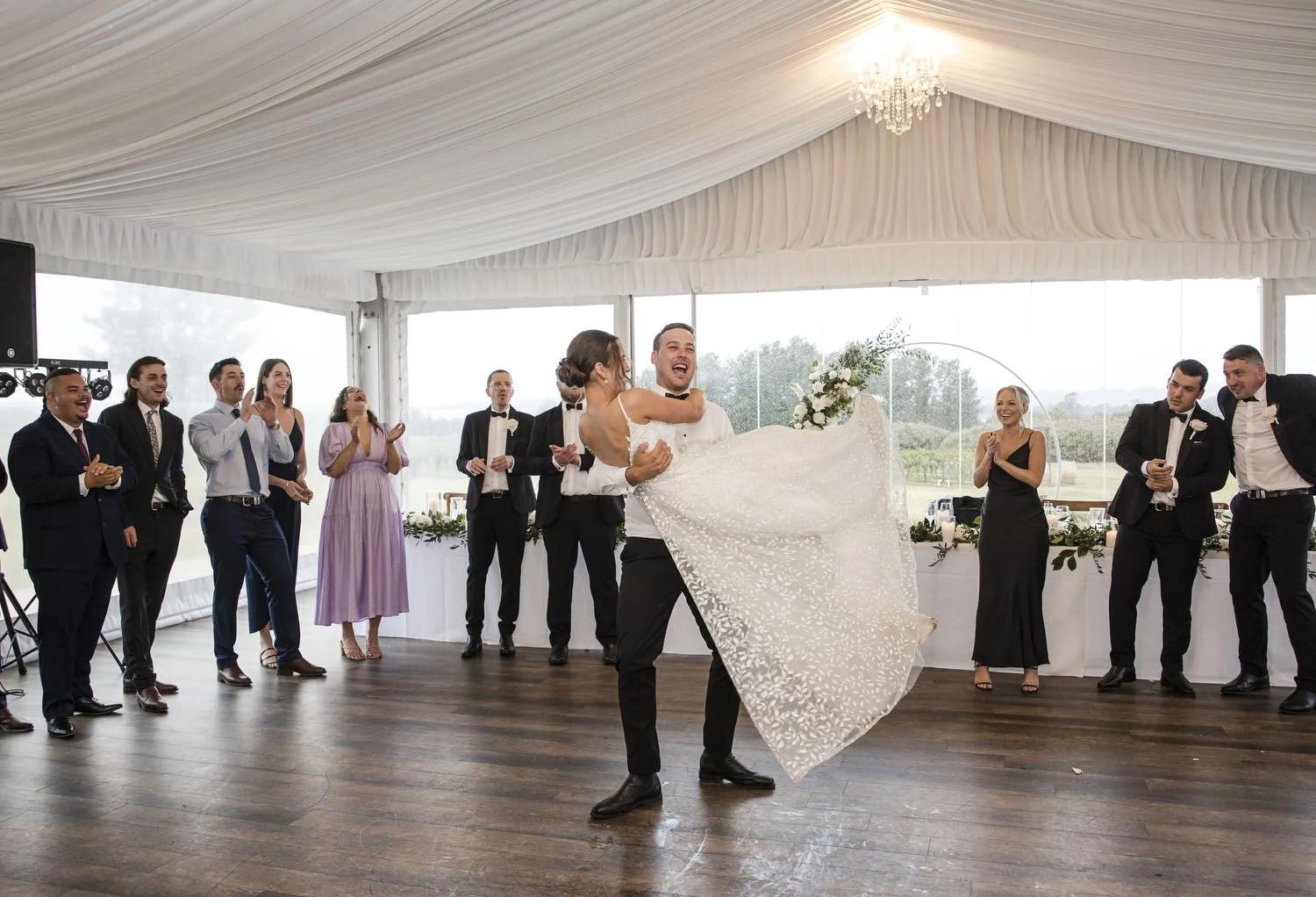 A couple dancing at a wedding reception in a tent with onlookers clapping and cheering.