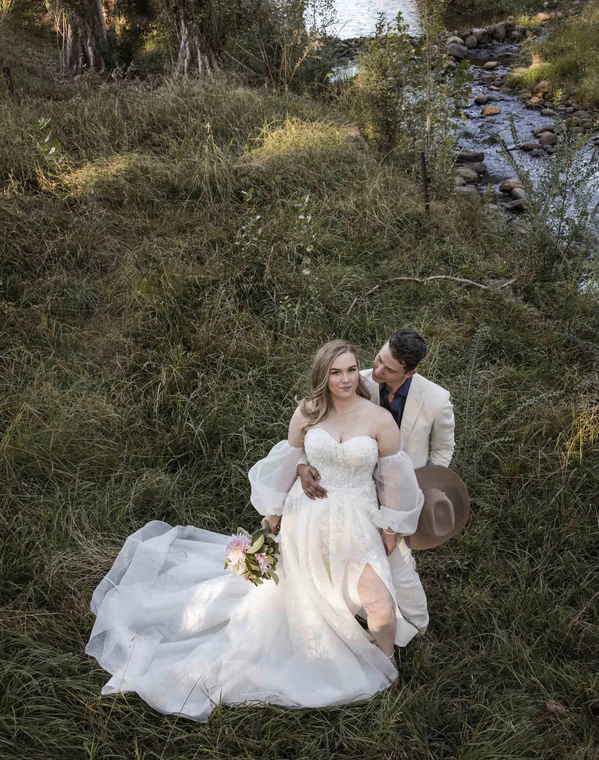 Bride and groom sitting together in long grass beside a rocky creek, bouquet in her lap and his hat in hand