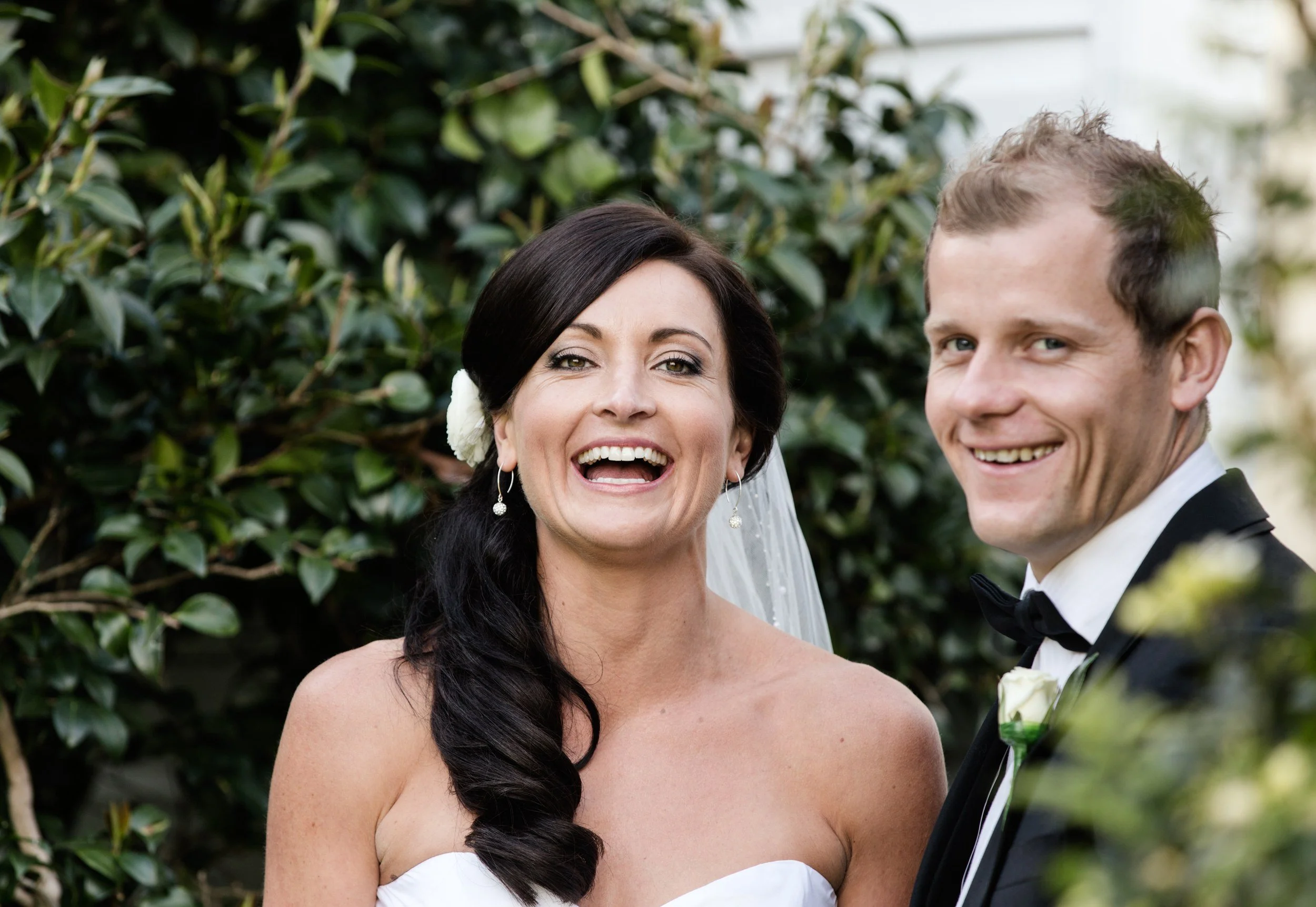 Bride and groom laughing together in front of a green hedge