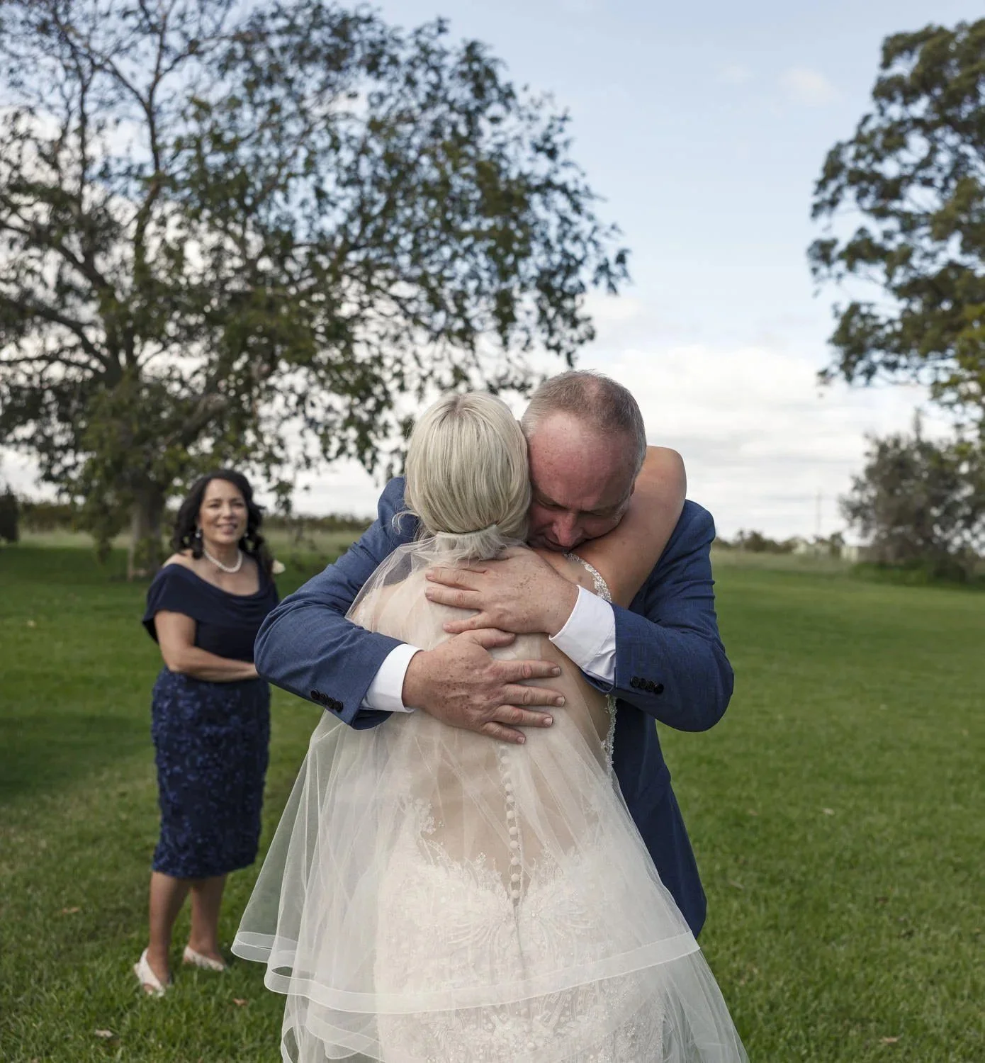 A man in a navy suit hugging the bride in her gown and veil on a lawn while a woman in navy smiles in the background