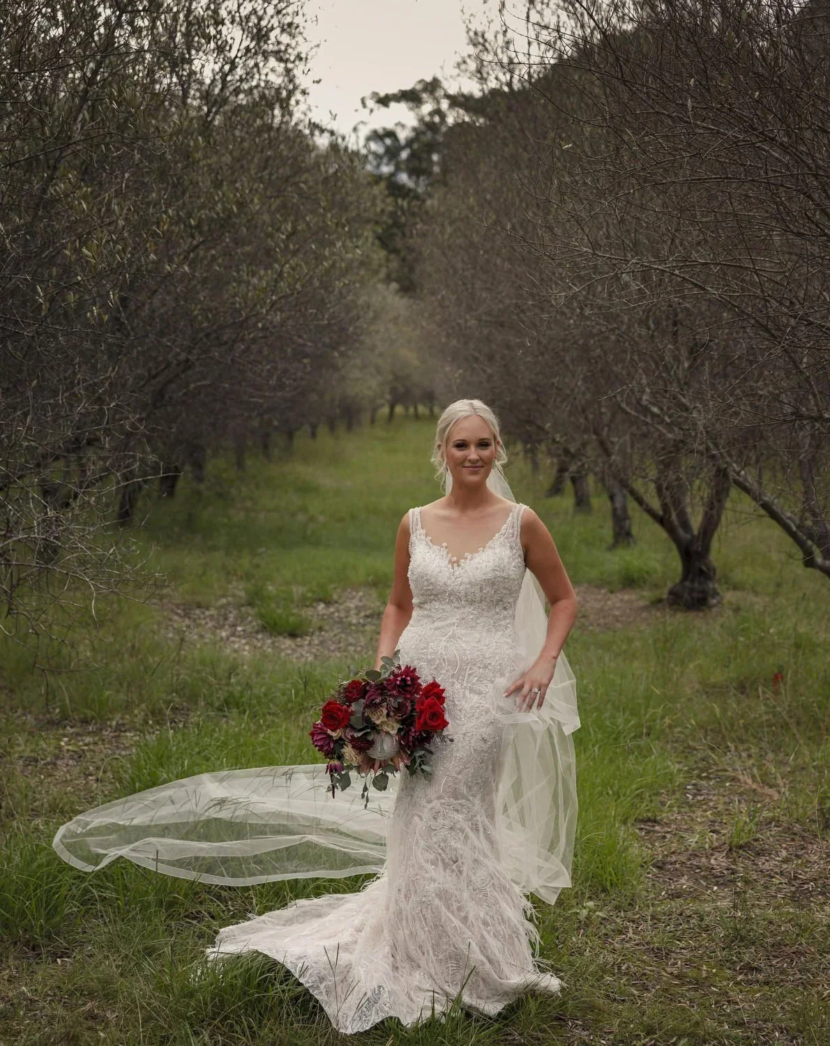 A bride holding a bouquet of red roses as her veil trails behind her in a row of trees