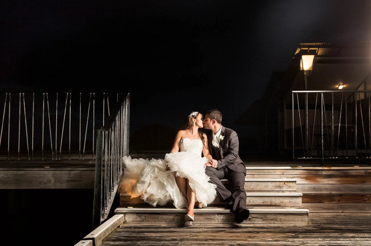 Bride and groom sitting on outdoor steps at night, kissing under a streetlight.