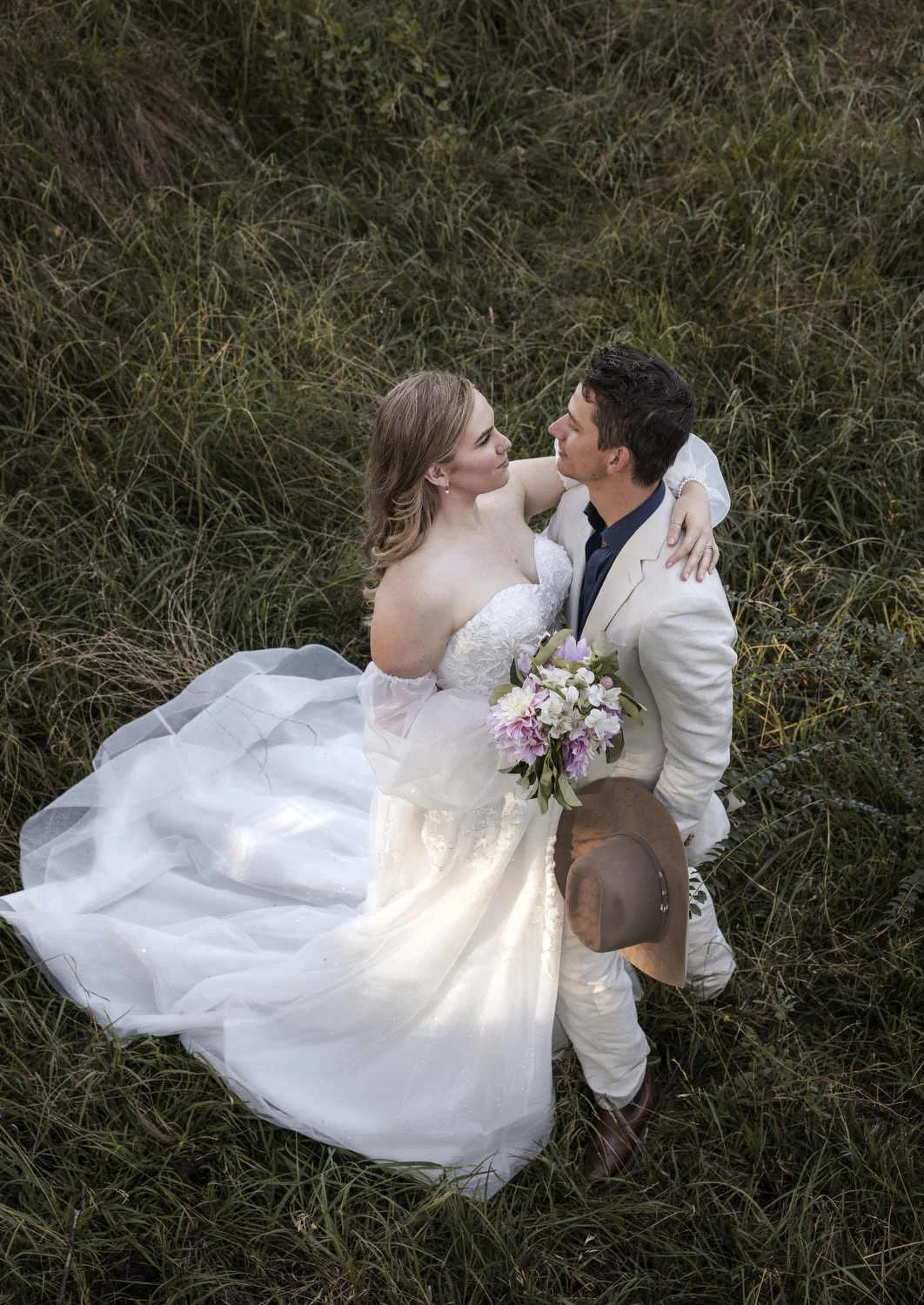 Couple embracing in tall grass, seen from above, with a pink and white bouquet and a felt hat