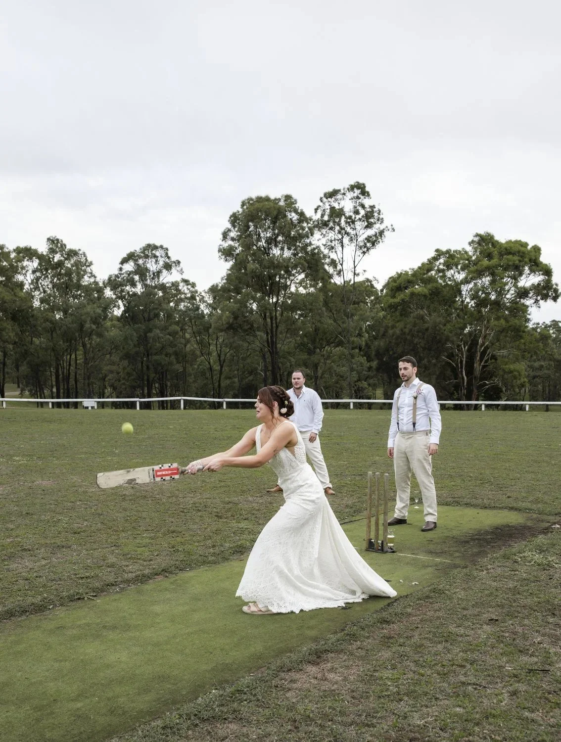 A bride in her wedding dress swinging a cricket bat at a ball on the pitch while two groomsmen watch