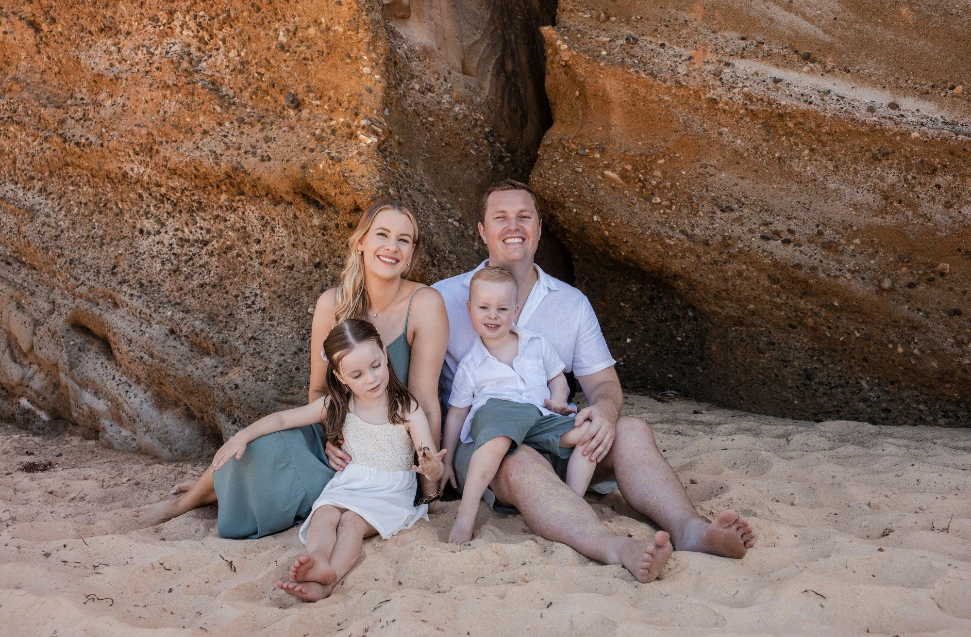 Family sitting together on the sand in front of an orange rock wall
