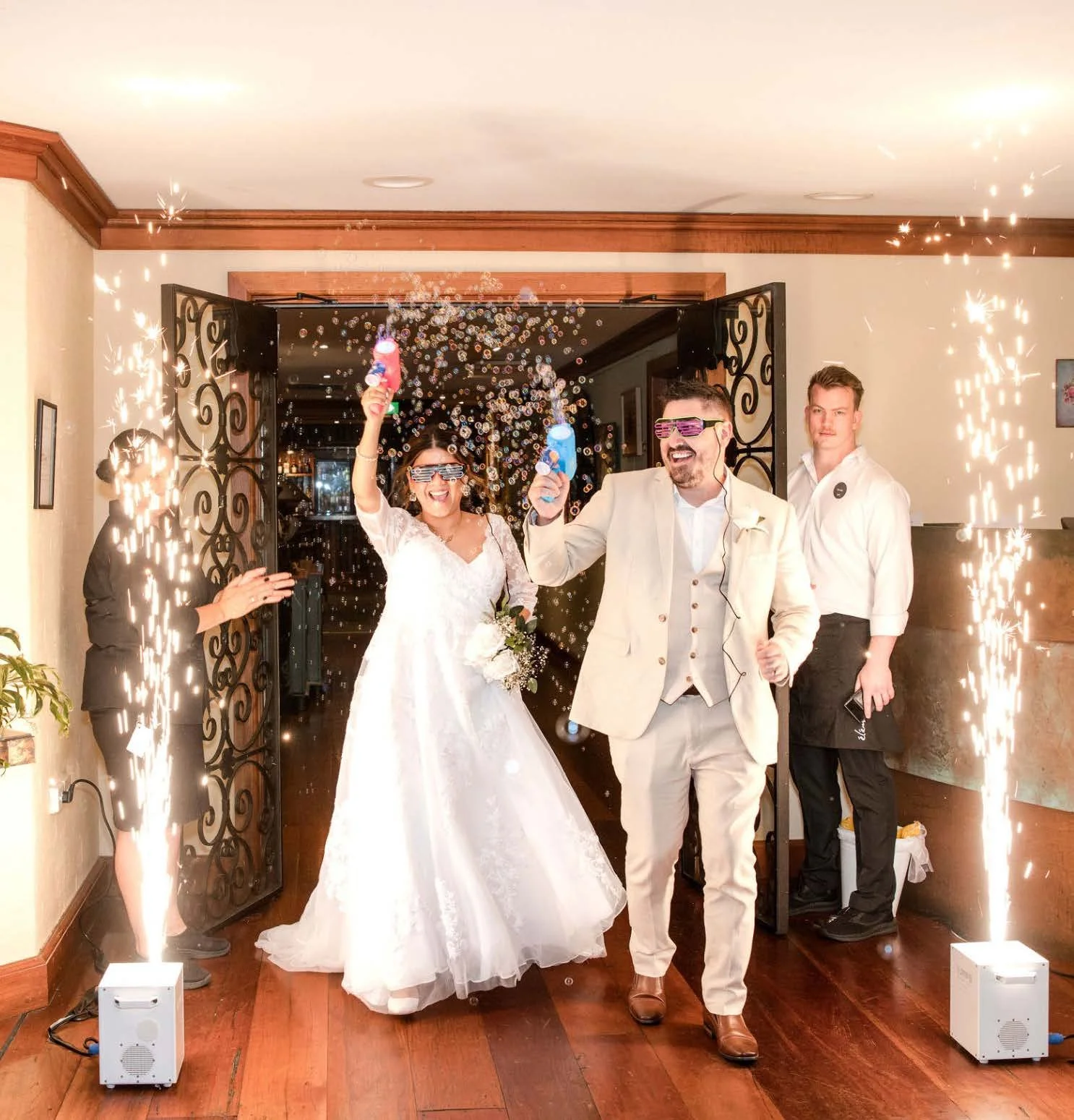 Bride and groom exiting a venue with bubble guns during a celebration, surrounded by sparklers and attendees.