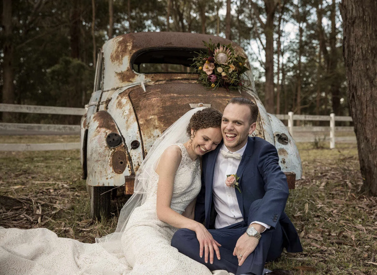 Bride and groom sitting in front of a rusty vintage car with flowers on its back, smiling, surrounded by trees.