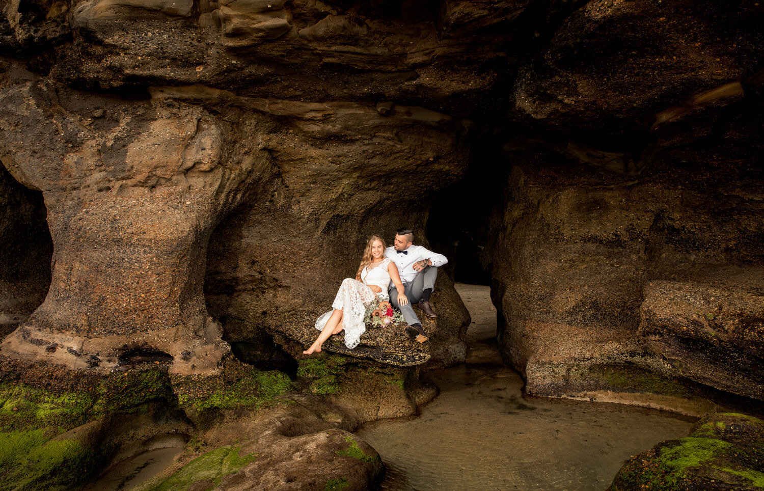 A couple sitting on a rock formation in a coastal cave, with the woman in a white dress and the man in a white shirt and gray pants, surrounded by moss and water.