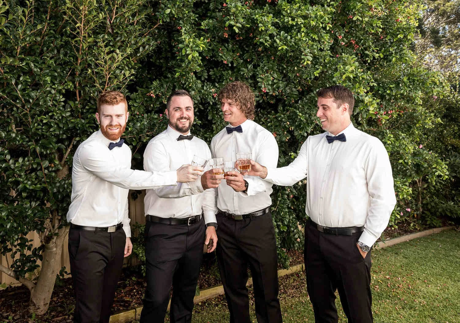 Four men dressed in formal white shirts and black bow ties, standing outdoors, clinking glasses, smiling.