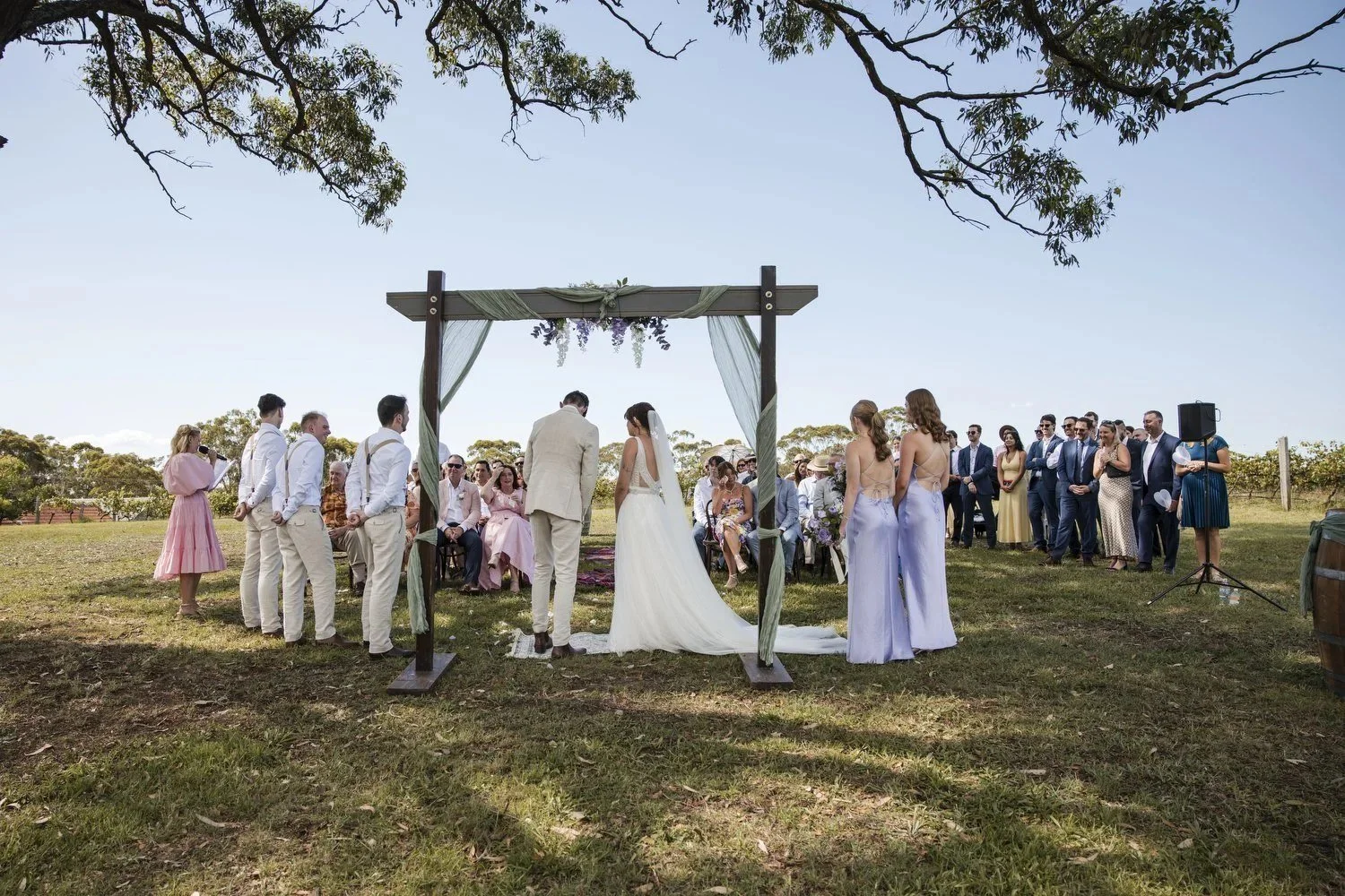 A wedding ceremony under a large gum tree at Wandin Valley Estate in Lovedale, with a timber arbour, bridesmaids in lilac and guests seated in front of the vineyard
