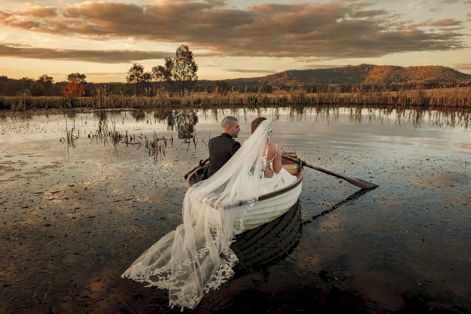 Bride and groom sitting in a rowboat on a lake at sunset, with a veil trailing in the water, surrounded by reeds and distant hills.