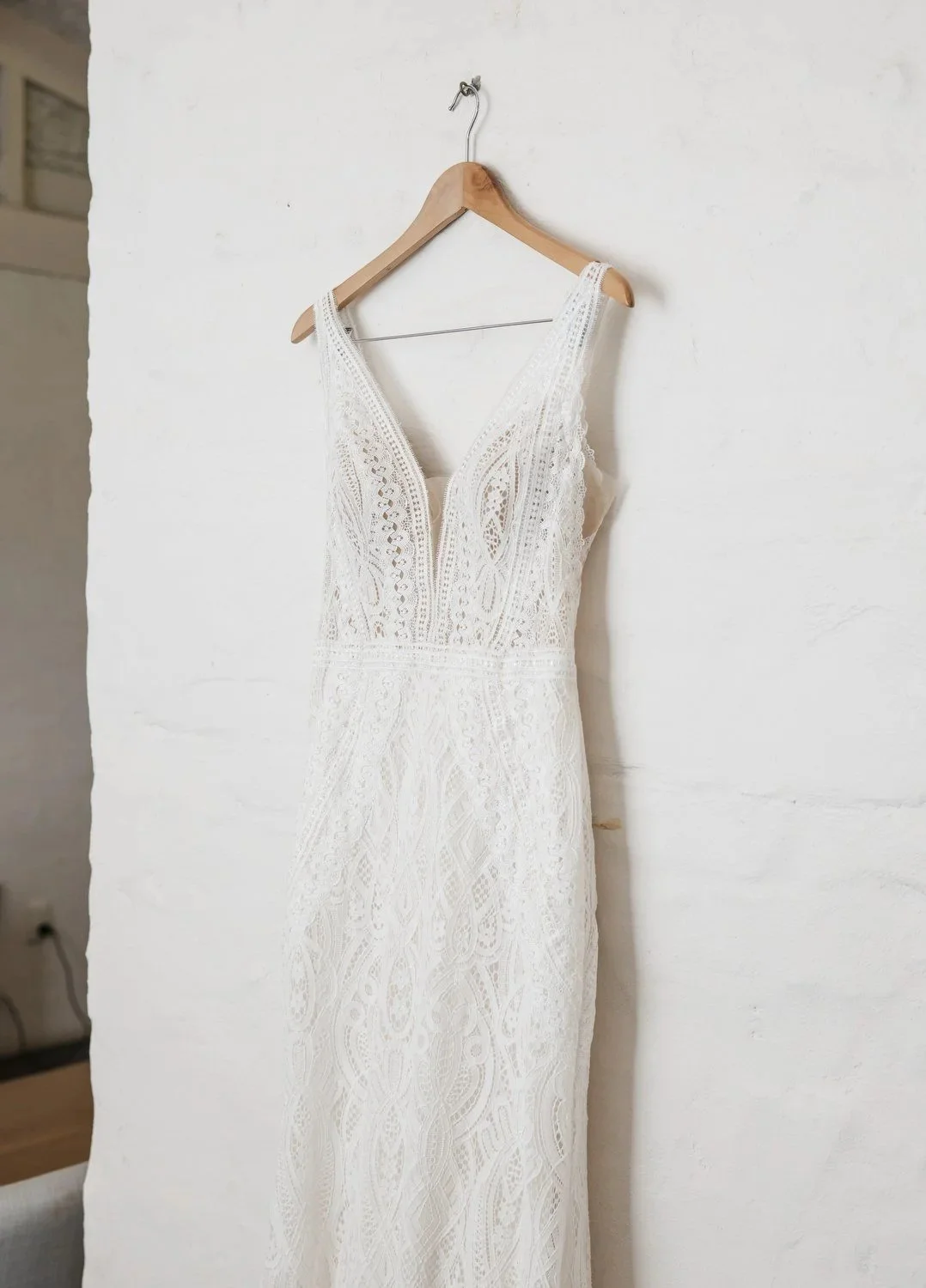 A white lace wedding dress hanging on a wooden hanger against a white brick wall