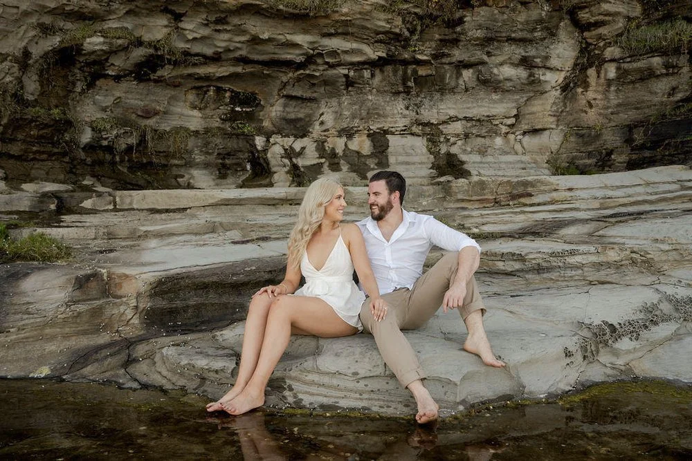 Couple sitting on rocky shoreline, smiling at each other, wearing casual beach attire.