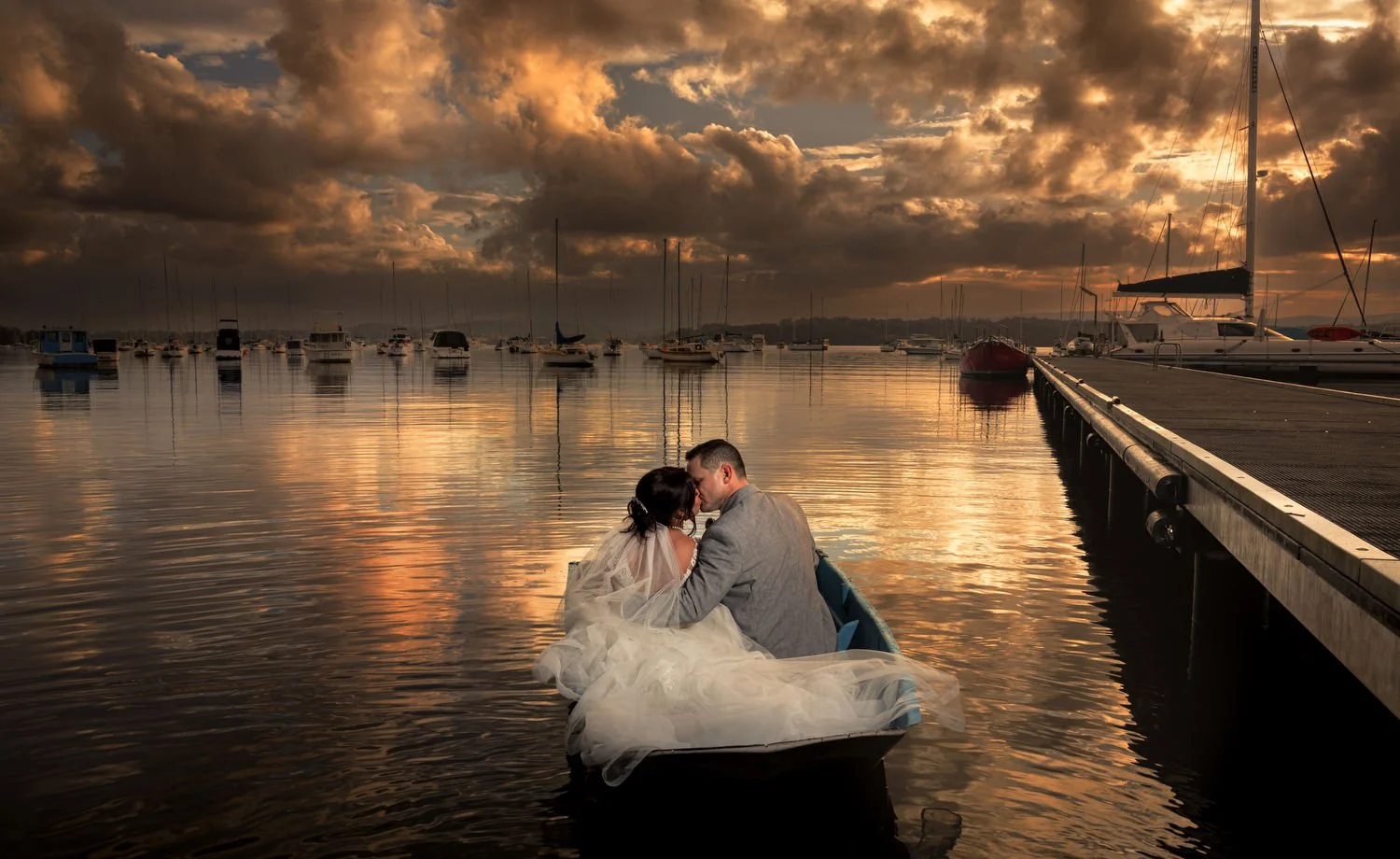 Bride and groom in a rowboat on a lake at sunset, surrounded by sailboats, with dramatic clouds in the sky.