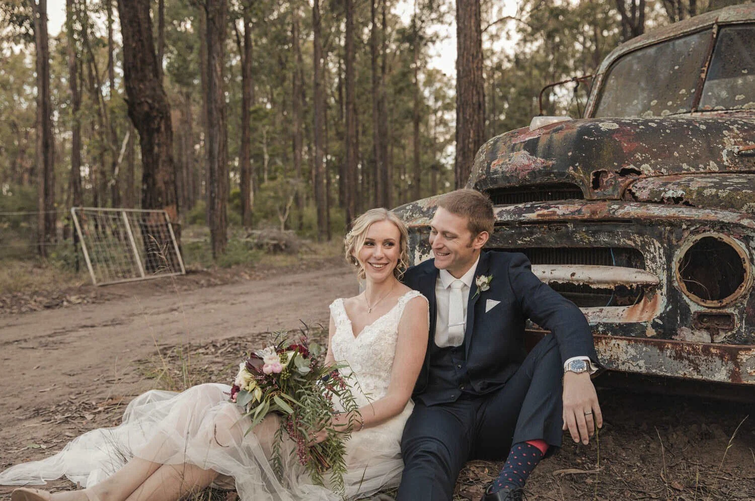 Bride and groom sitting by a rustic truck in a forest setting.