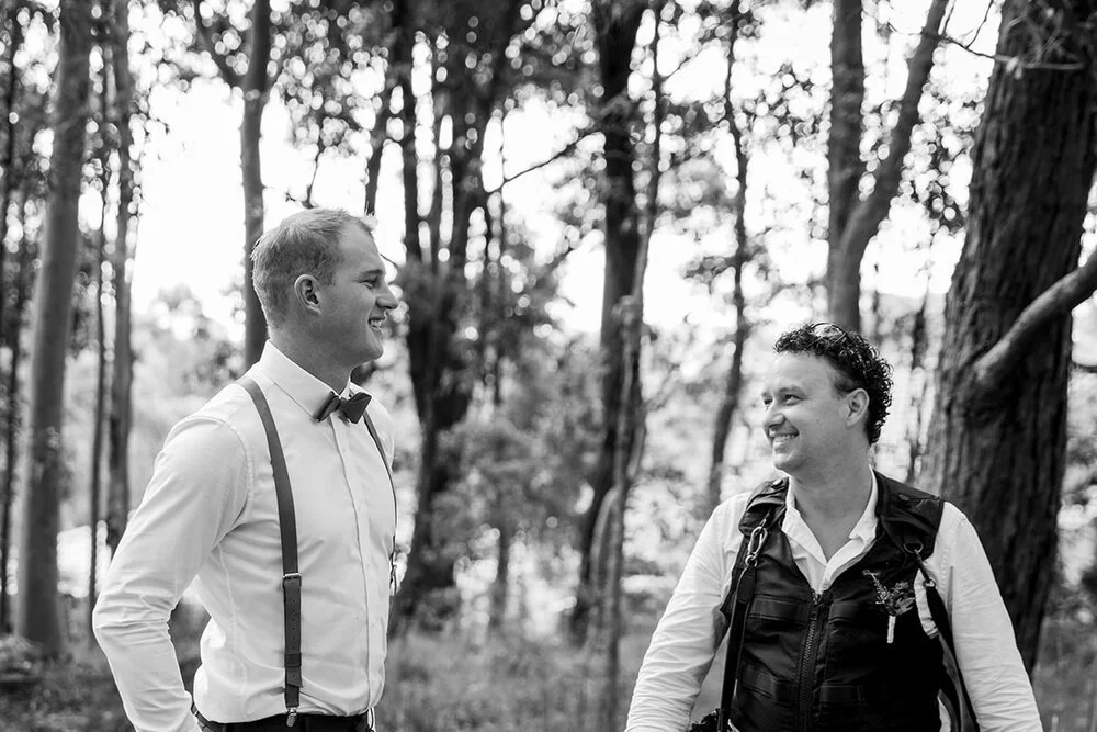 Two men smiling at each other in a forest, one wearing suspenders and a bowtie, and the other wearing a vest.