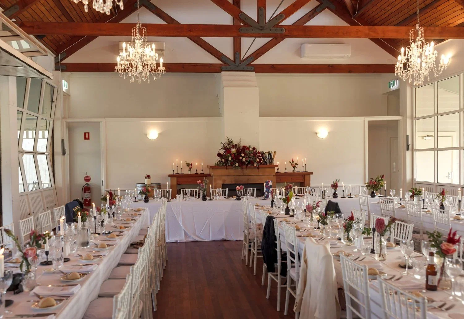 The Cricketer's Pavilion set for a wedding reception, with long tables, candles, red flowers, chandeliers and exposed timber beams