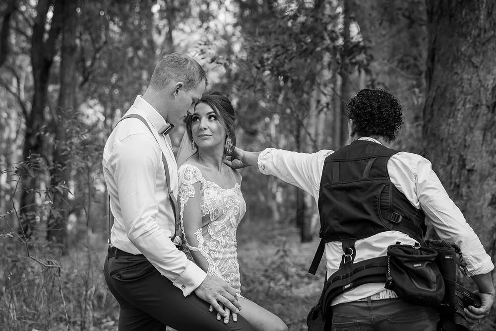 Black and white photo of a couple in wedding attire posing in a forest with a photographer adjusting the bride's position.