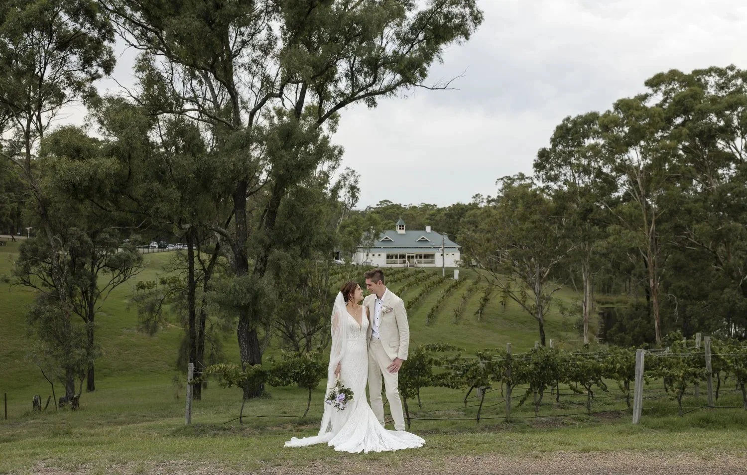 A bride and groom standing close in front of the vineyard, with gum trees and a white estate building on the hill behind them