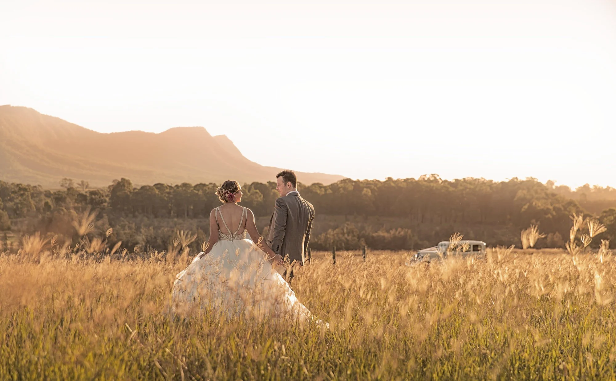 Bride and groom walking through a grassy field at sunset with mountains in the background.