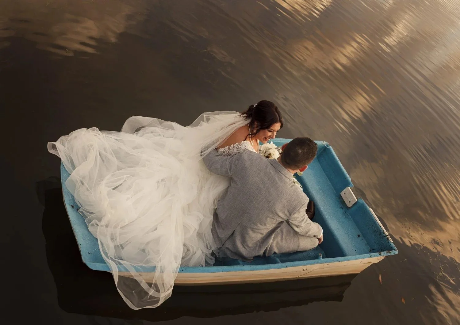 Bride and groom cuddling in a blue boat on dark water, the veil draped over the side