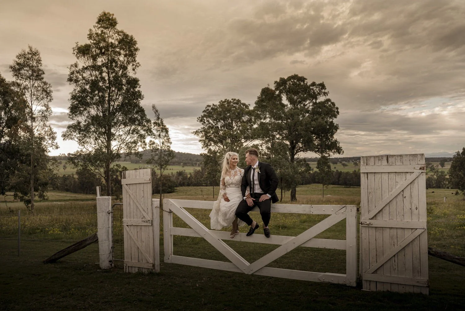 Wedding couple sitting on a white farm gate with a rural landscape and trees in the background, under a cloudy sky.