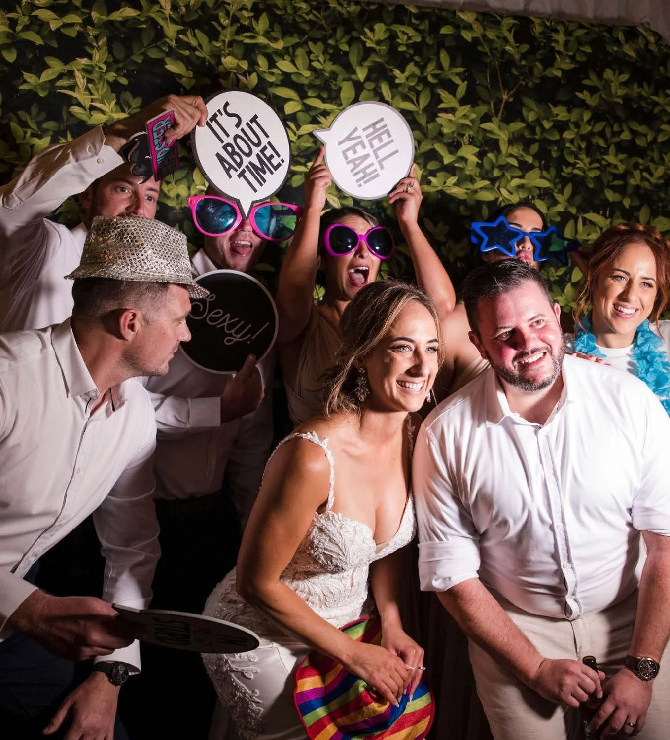 A group of people at a celebration, possibly a wedding reception, posing with humorous props and signs such as "It's About Time!" and "Hell Yeah!" in front of a leafy backdrop. They are smiling and wearing fun accessories like hats and oversized glasses.