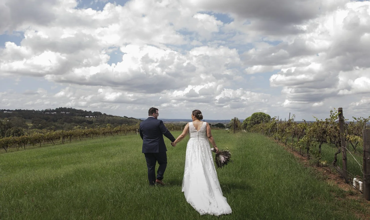 Couple walking hand in hand down a grassy vineyard avenue under a wide sky