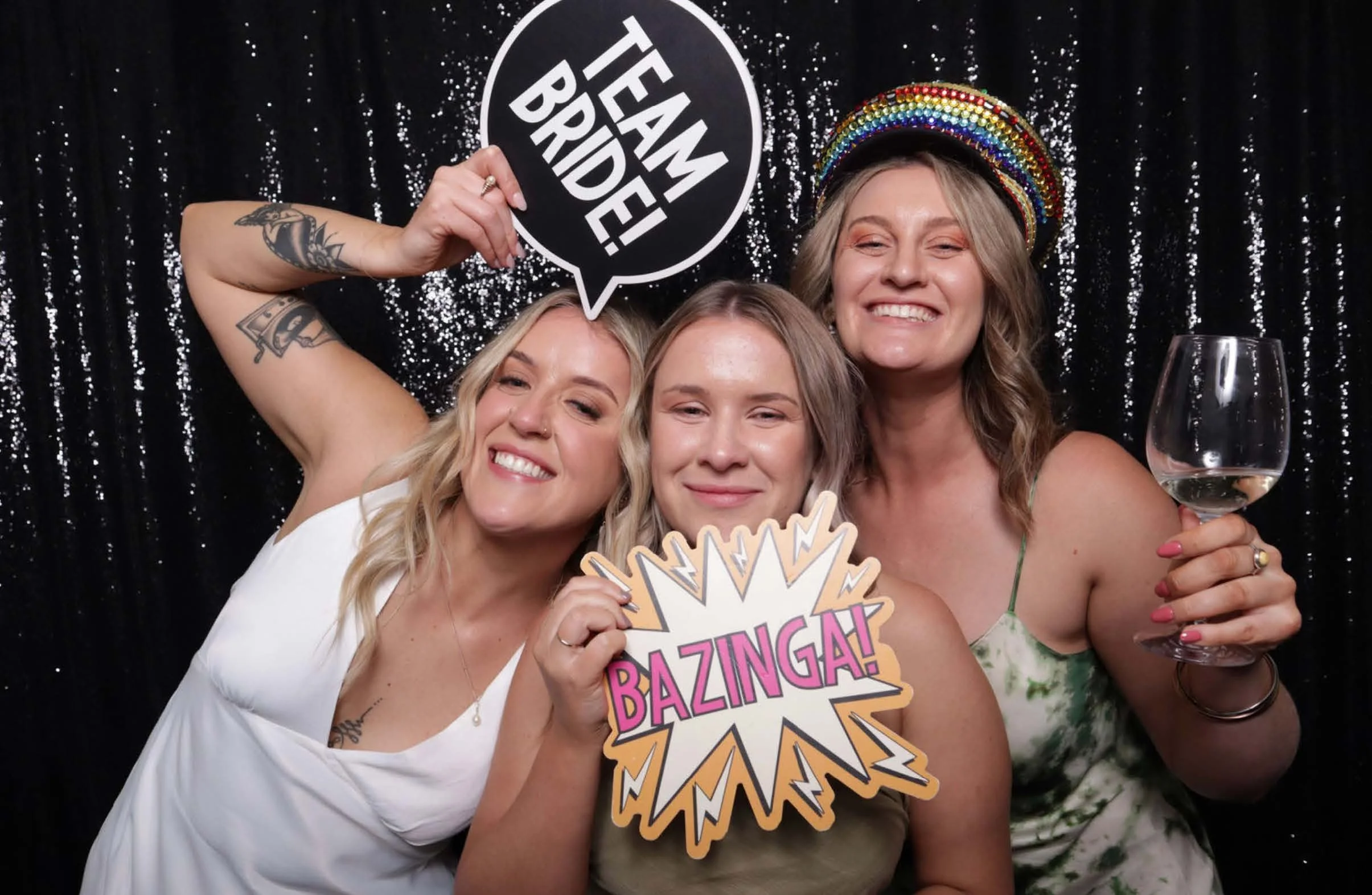 Three women posing at a photo booth with props, including a "Team Bride!" sign and a "Bazinga!" sign. One woman holds a glass of white wine, wearing a colorful hat against a black sequin background.
