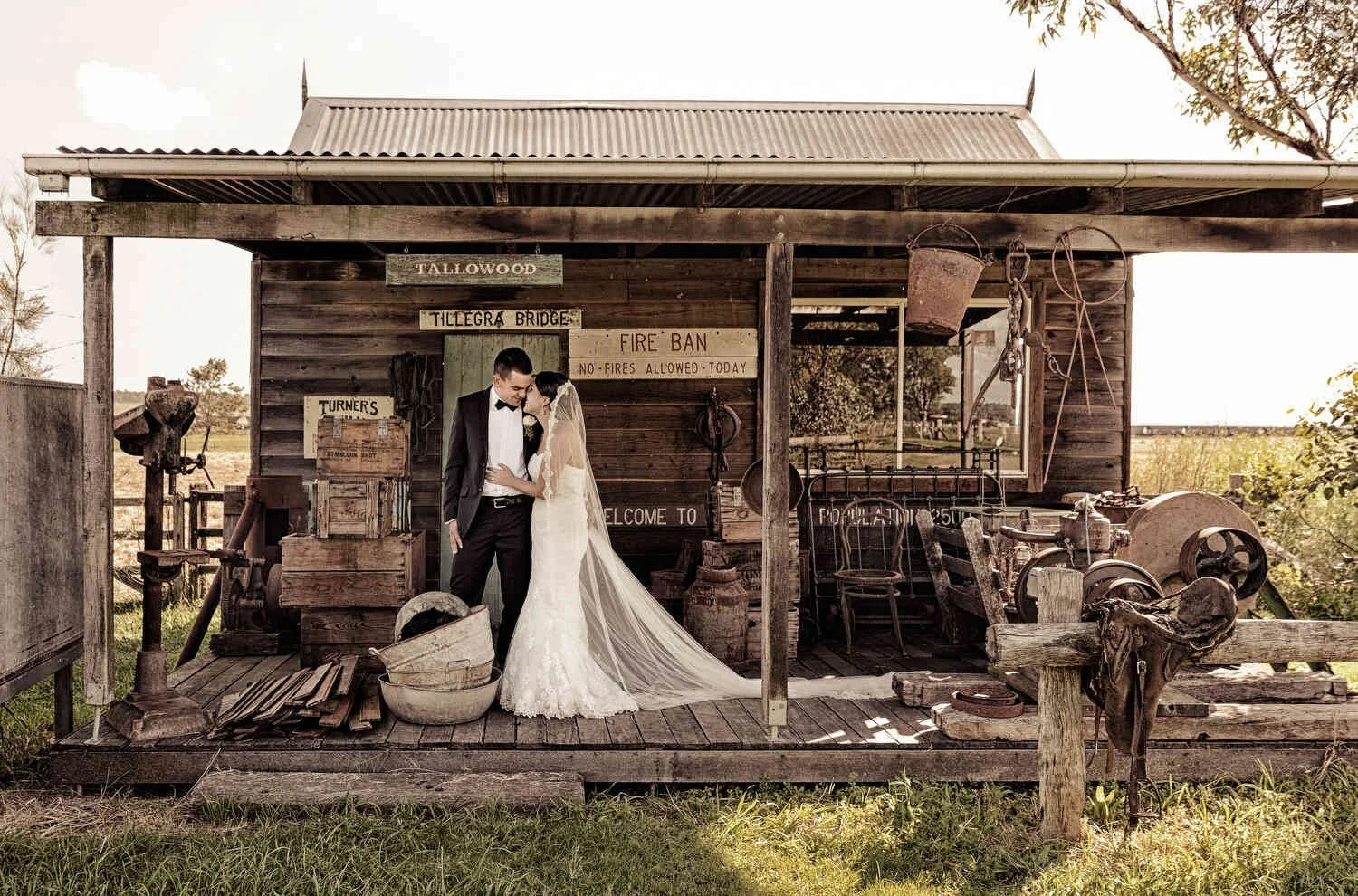 A bride and groom embrace on the porch of a rustic wooden cabin, surrounded by vintage signs and farm equipment, in a rural setting.
