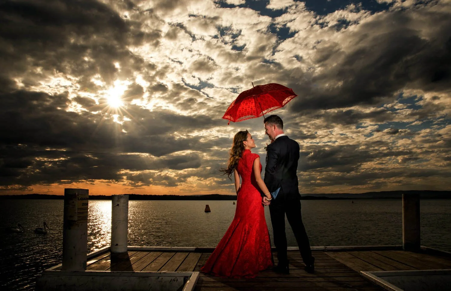 Couple on a pier during sunset holding a red umbrella, with a dramatic cloudy sky and sunlight reflecting on the water.