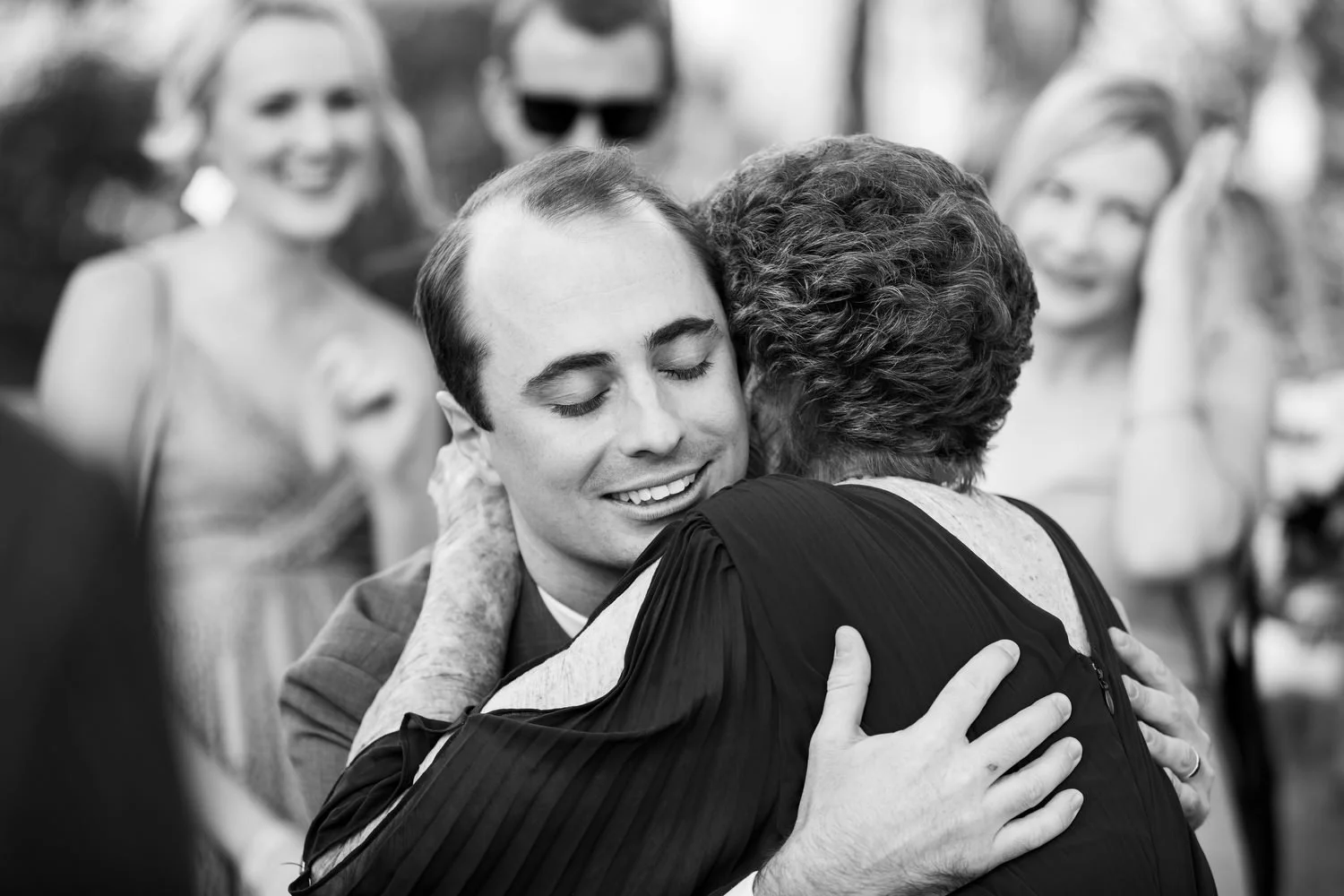 Black and white photo of a man hugging an elderly woman at an event, with guests smiling in the background.