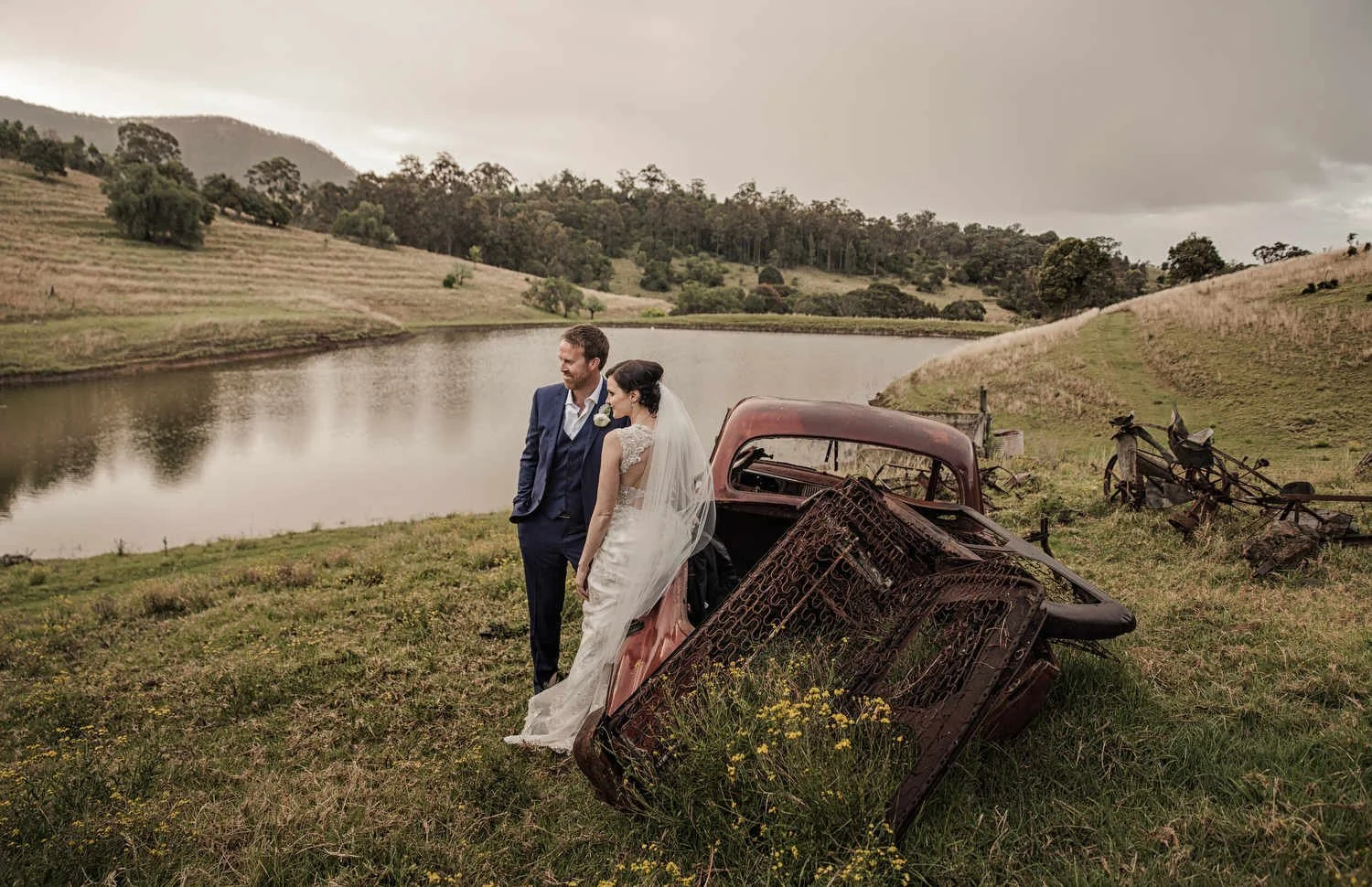 Bride and groom standing near a lake with rusted car parts on grassy hill