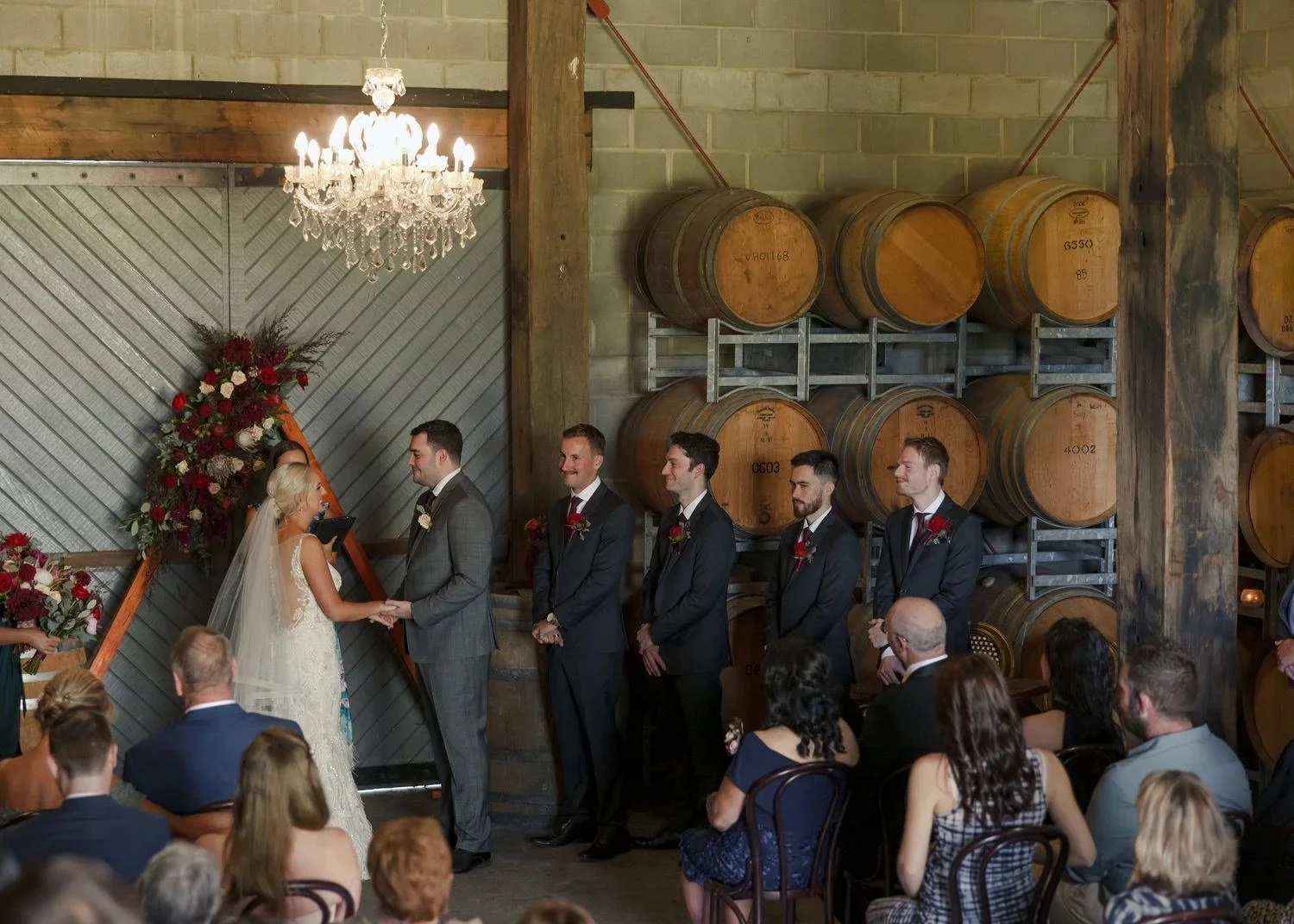 A couple holding hands during their ceremony in a barn lined with oak wine barrels, under a crystal chandelier