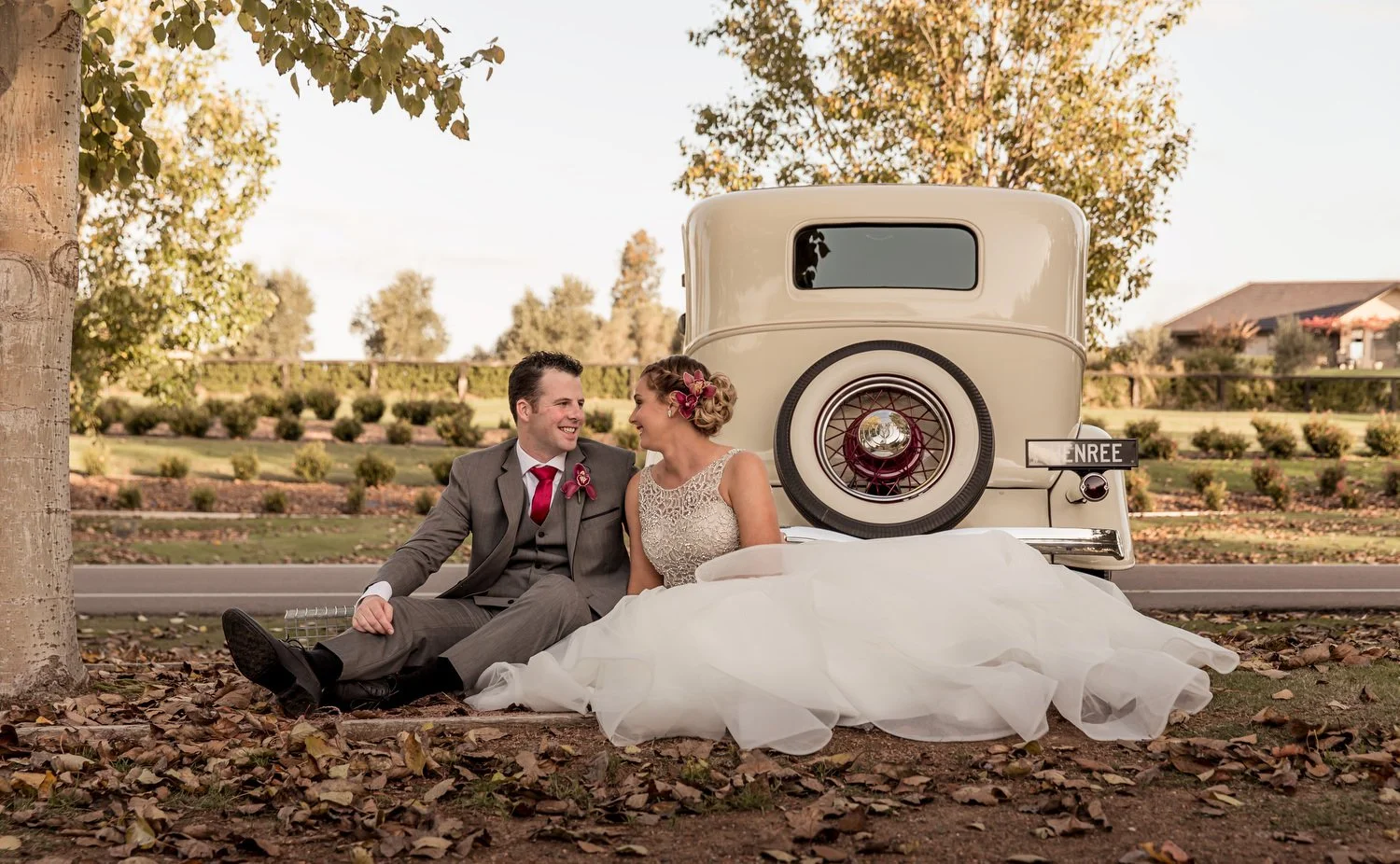 Bride and groom sitting on the ground, smiling at each other in wedding attire, with a vintage car behind them, surrounded by trees and fallen leaves.
