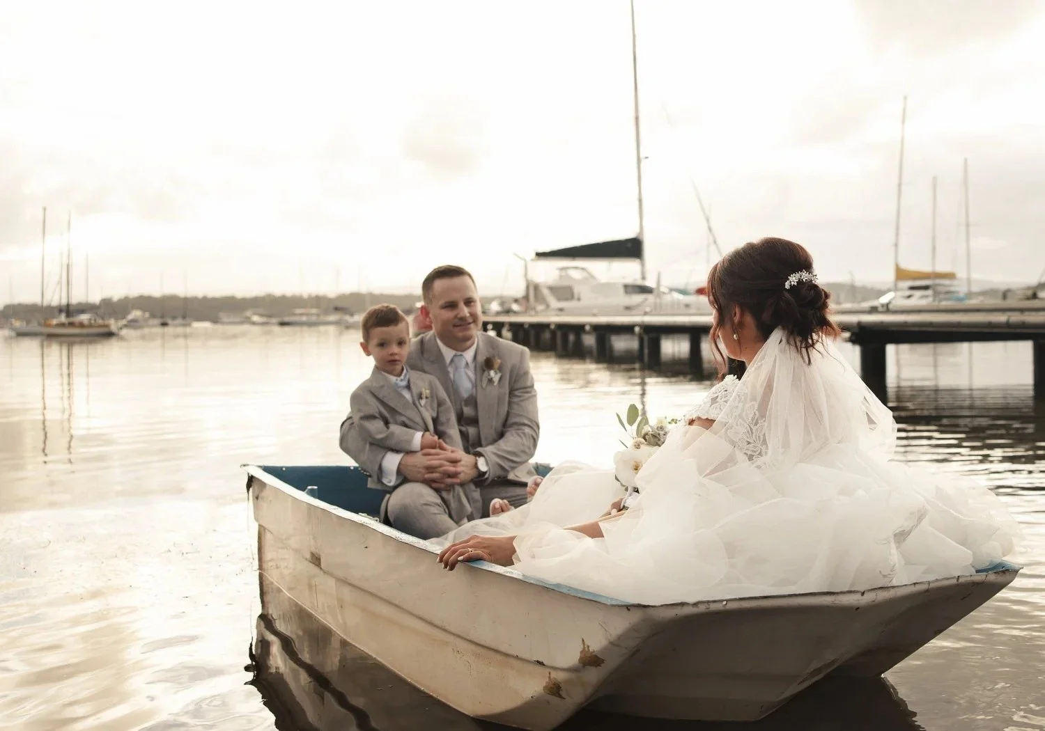 Bride and groom sitting in a small boat at the water's edge with yachts behind