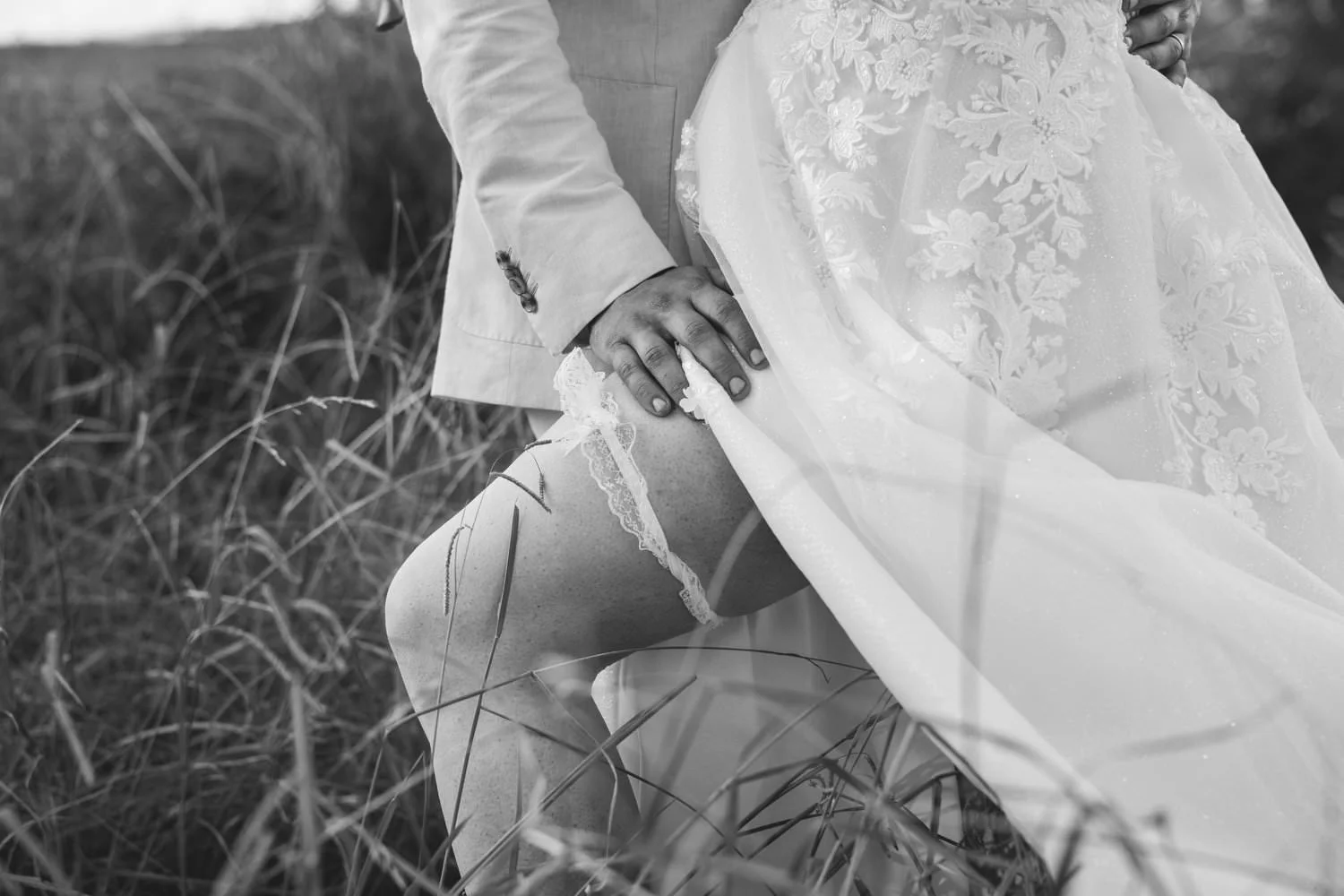 Black and white close-up of a groom's hand on the lace garter beneath a flowing wedding dress in long grass