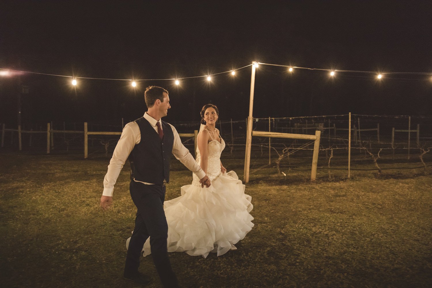 Bride and groom walking hand in hand outdoors at night under string lights.