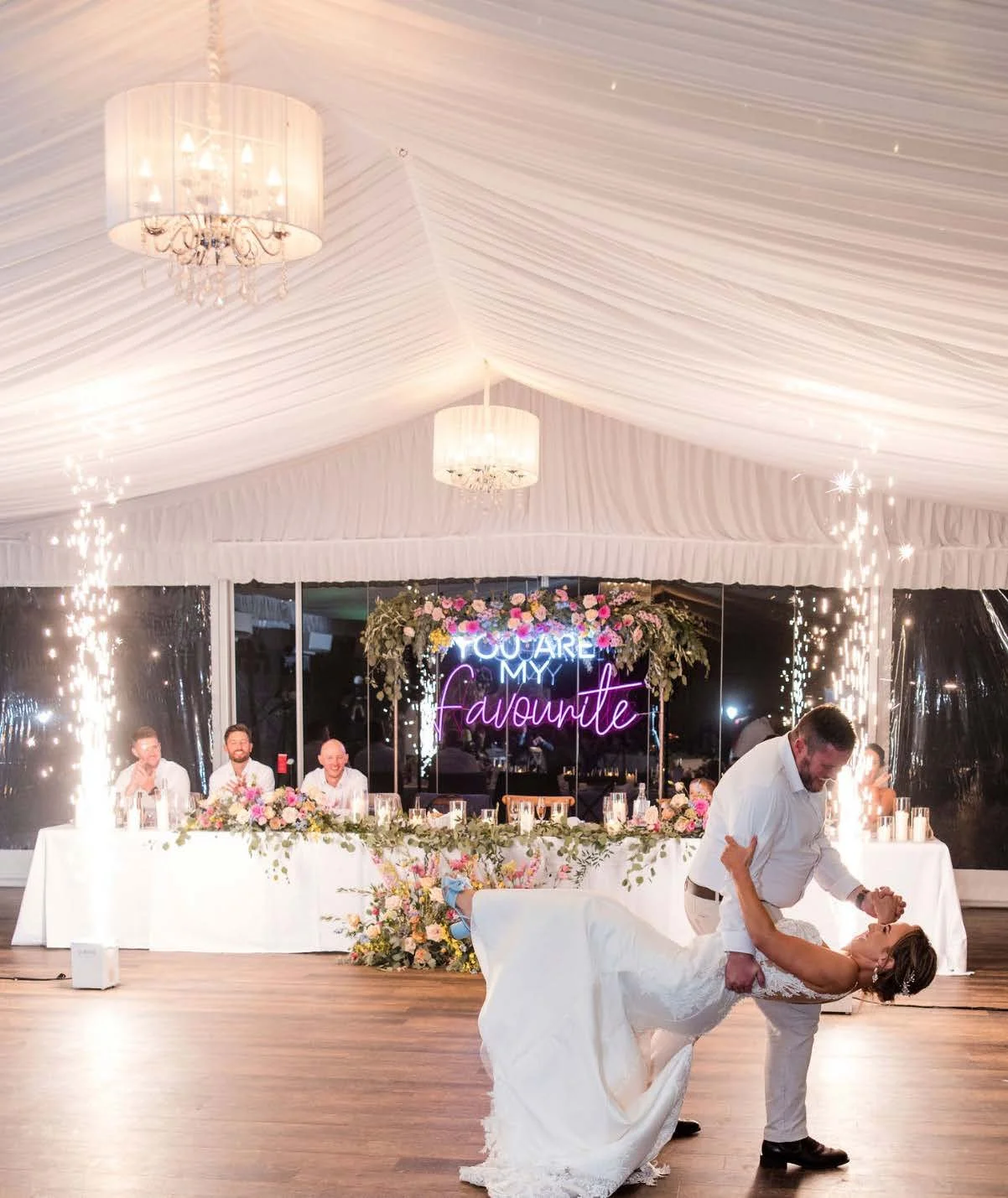 A wedding reception with a couple dancing. The background includes a sign reading "You Are My Favorite," floral decorations, sparklers, and a long table with seated guests.