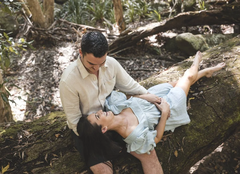 Couple sitting on a fallen tree in a forest, smiling at each other.