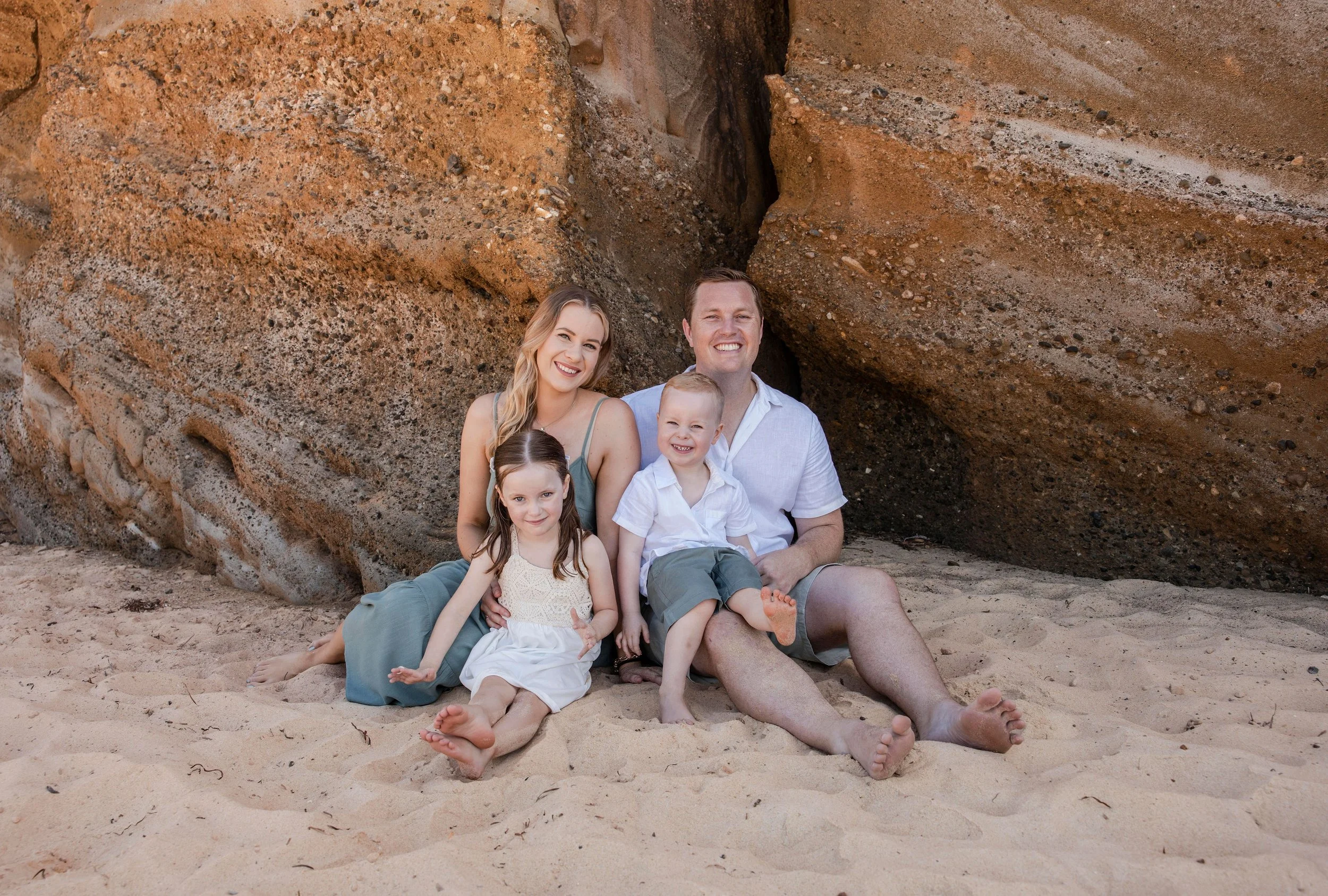 Family of four sitting close together on the sand beneath large sandstone rocks, smiling