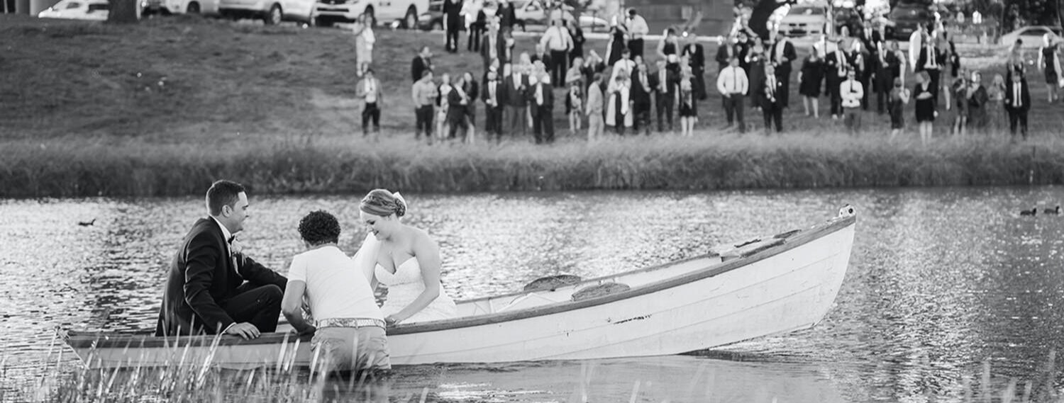 Bride and groom in a boat on a lake with wedding guests onshore in the background.